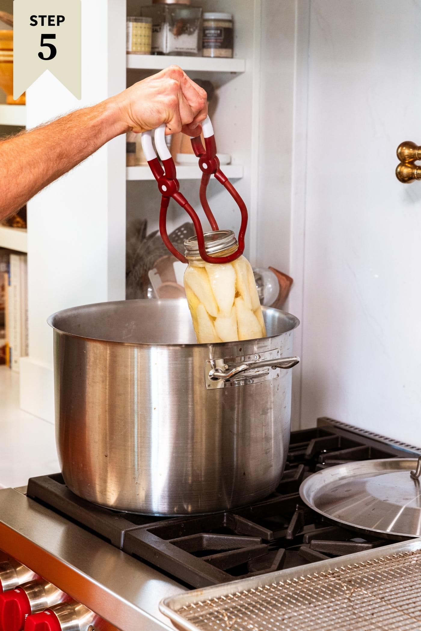 Hand removing glass canning jar filled with pears from metal kettle on stovetop.