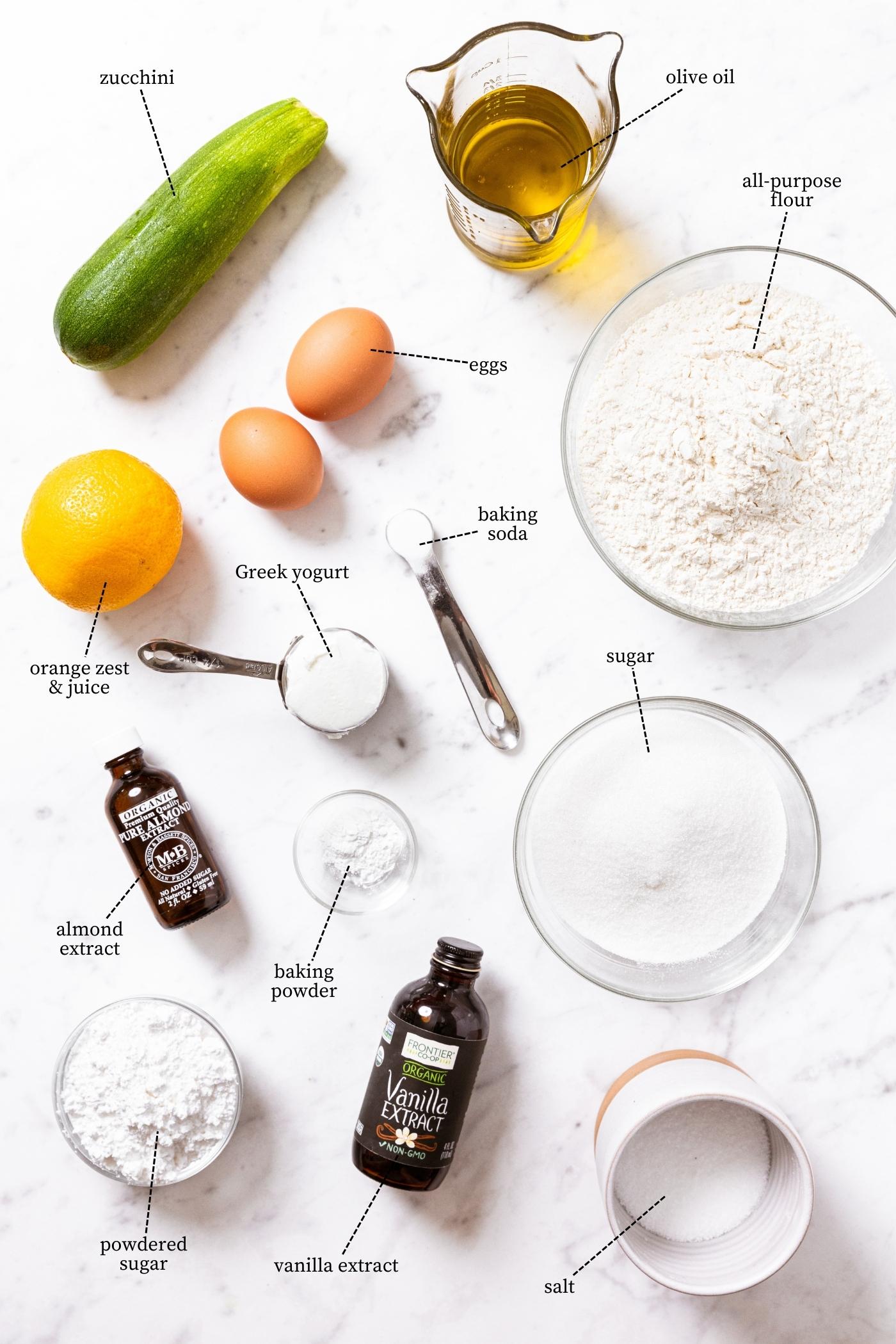 White marble surface holding all ingredients needed to make a zucchini olive oil cake including zucchini, olive oil, flour, sugar, orange, eggs, and more.