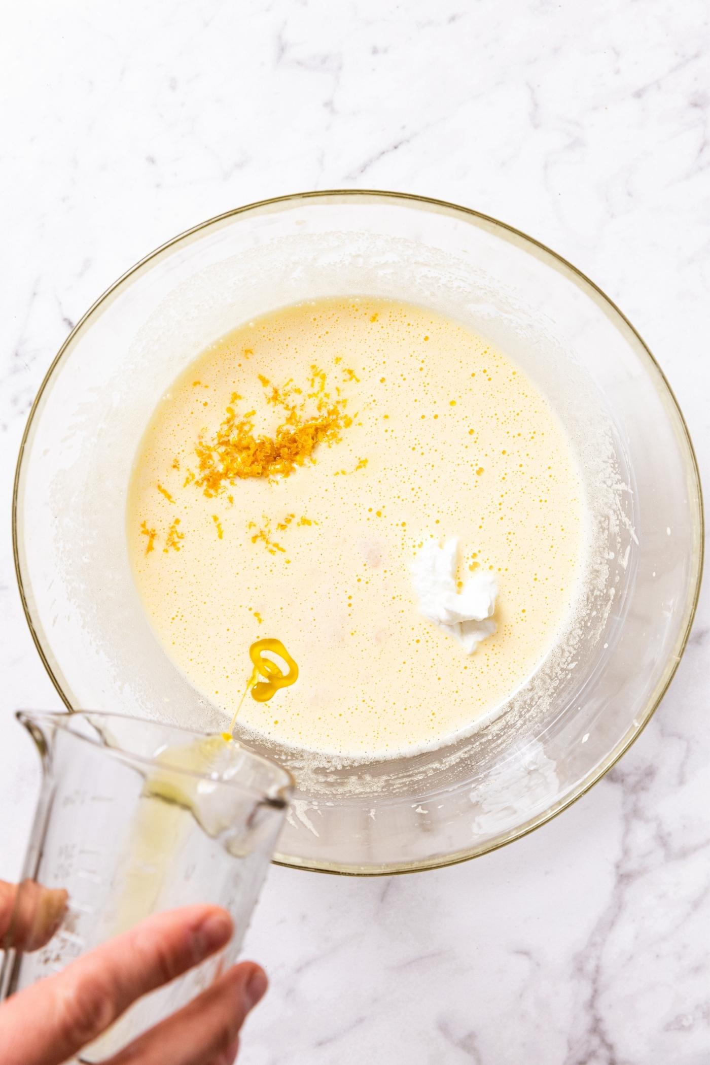 Glass bowl filled with wet batter with bits of ingredients showing with glass measuring cup pouring in last remaining amount of olive oil all on marble surface.