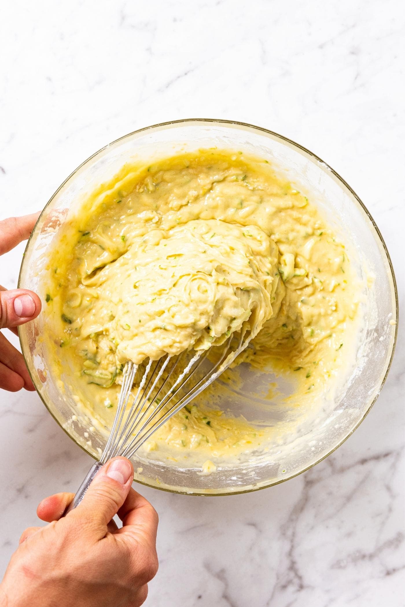Hand mixing together wet batter with metal whisk in glass bowl on marble countertop.