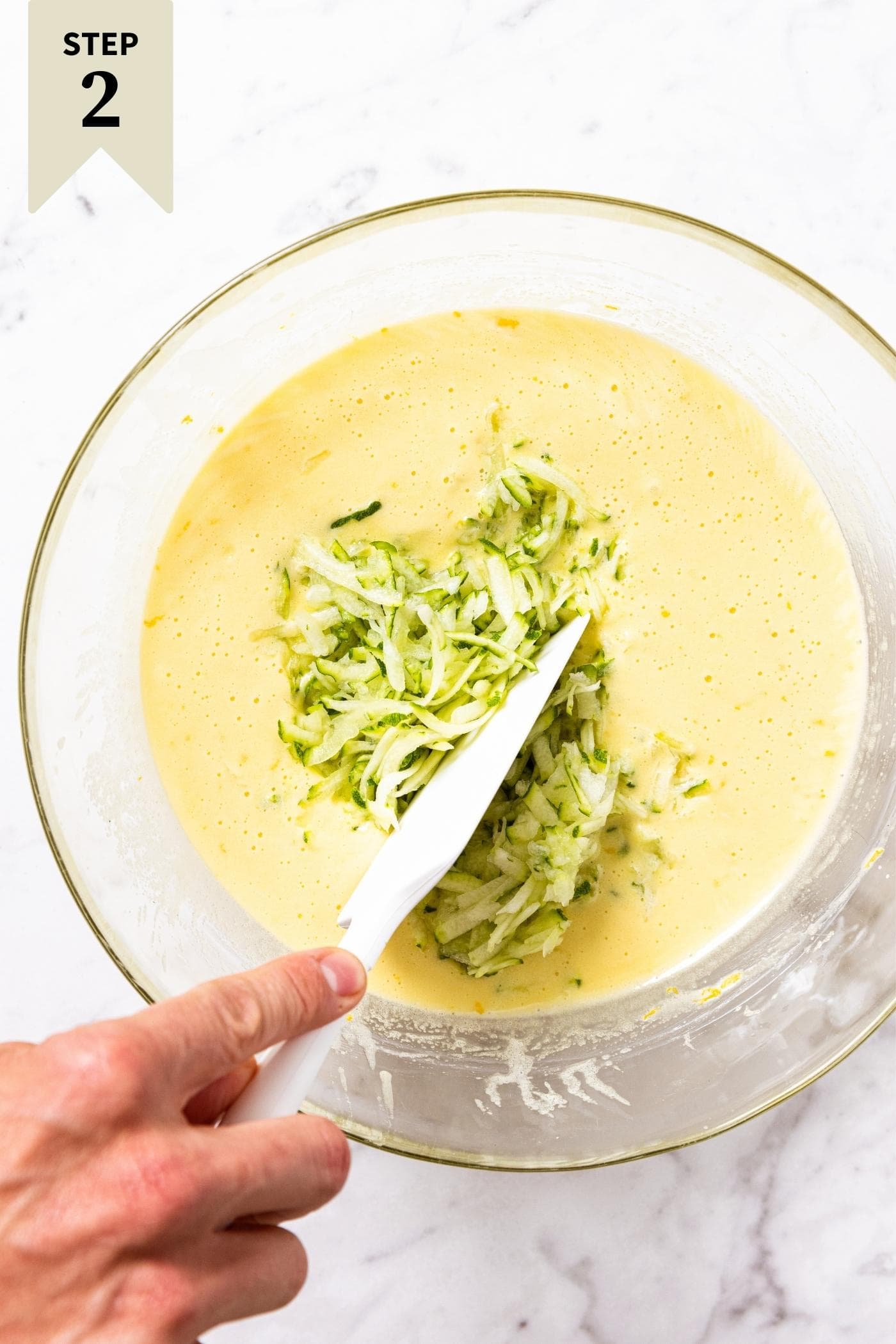 Hand about to mix up zucchini into yellow wet batter with white spatula all on marble surface.