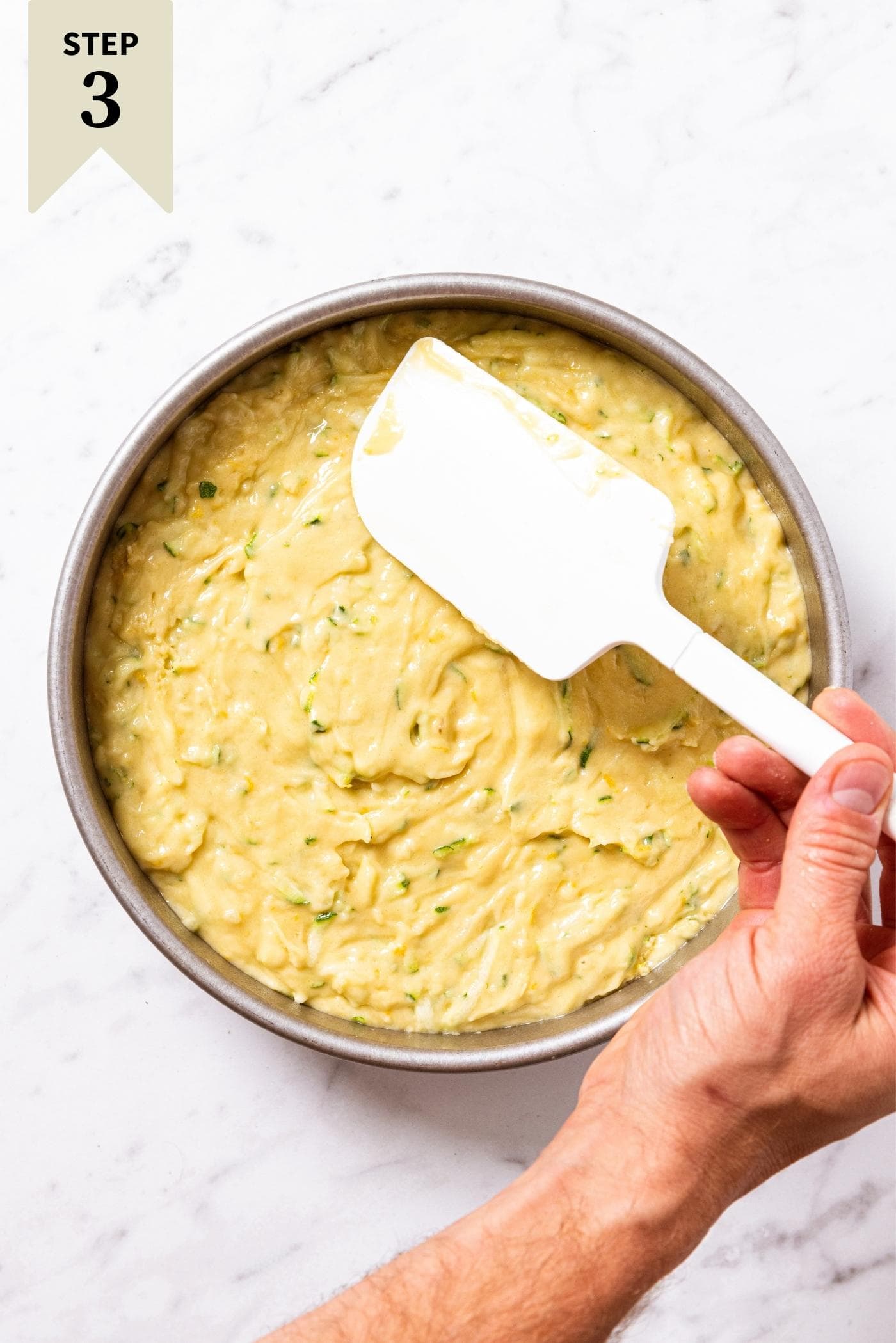 Hand smoothing out the top of batter in round cake pan with white spatula with the pan sitting on marble surface.