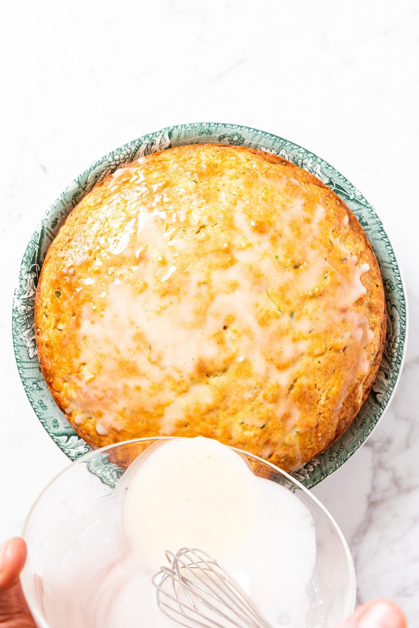 Hands holding glass bowl filled with remaining white colored glaze pouring on top of golden cake sitting on green cake plate on marble surface.