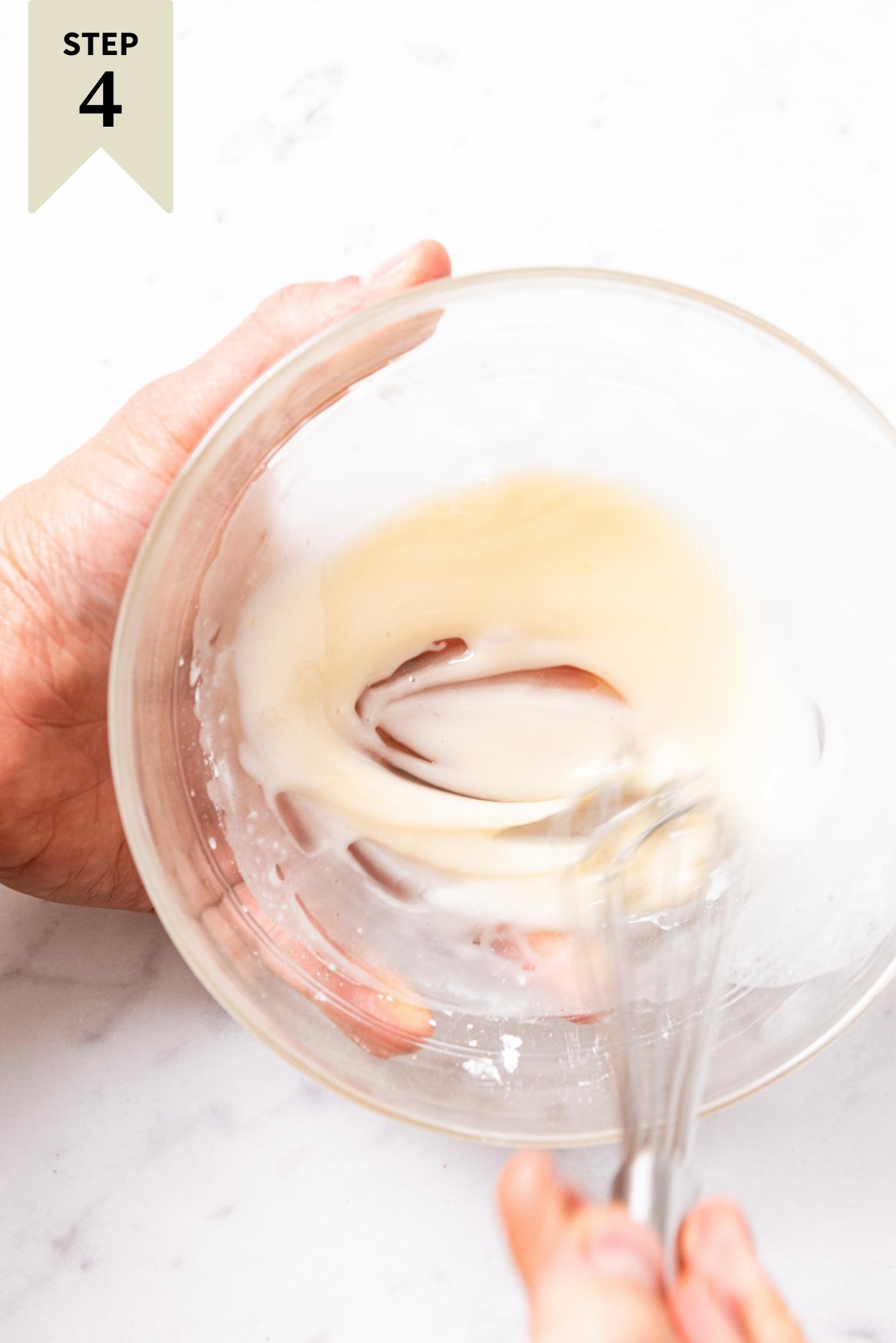 Hand holding glass bowl mixing together a small amount of light colored glaze that will be put on a cake.