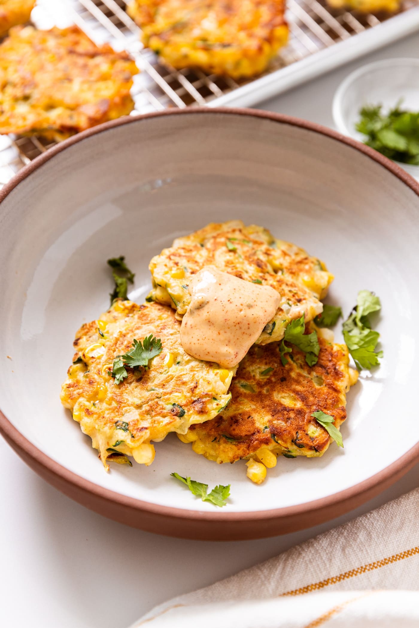White bowl filled with three fried zucchini and corn fritters topped with chipotle mayonnaise sauce with cilantro sprinkled on top with more fritters cooling in background.