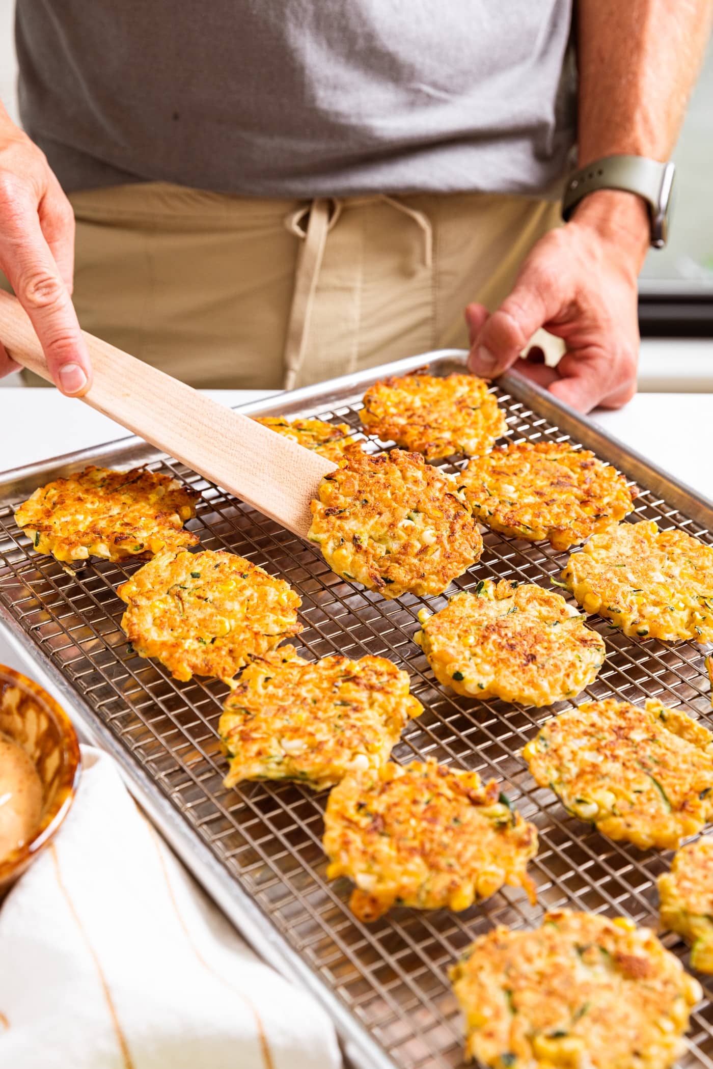 Hand holding wooden spatula scooping up one fried zucchini and corn fritter off of metal cooling rack filled with fritters all on white countertop.
