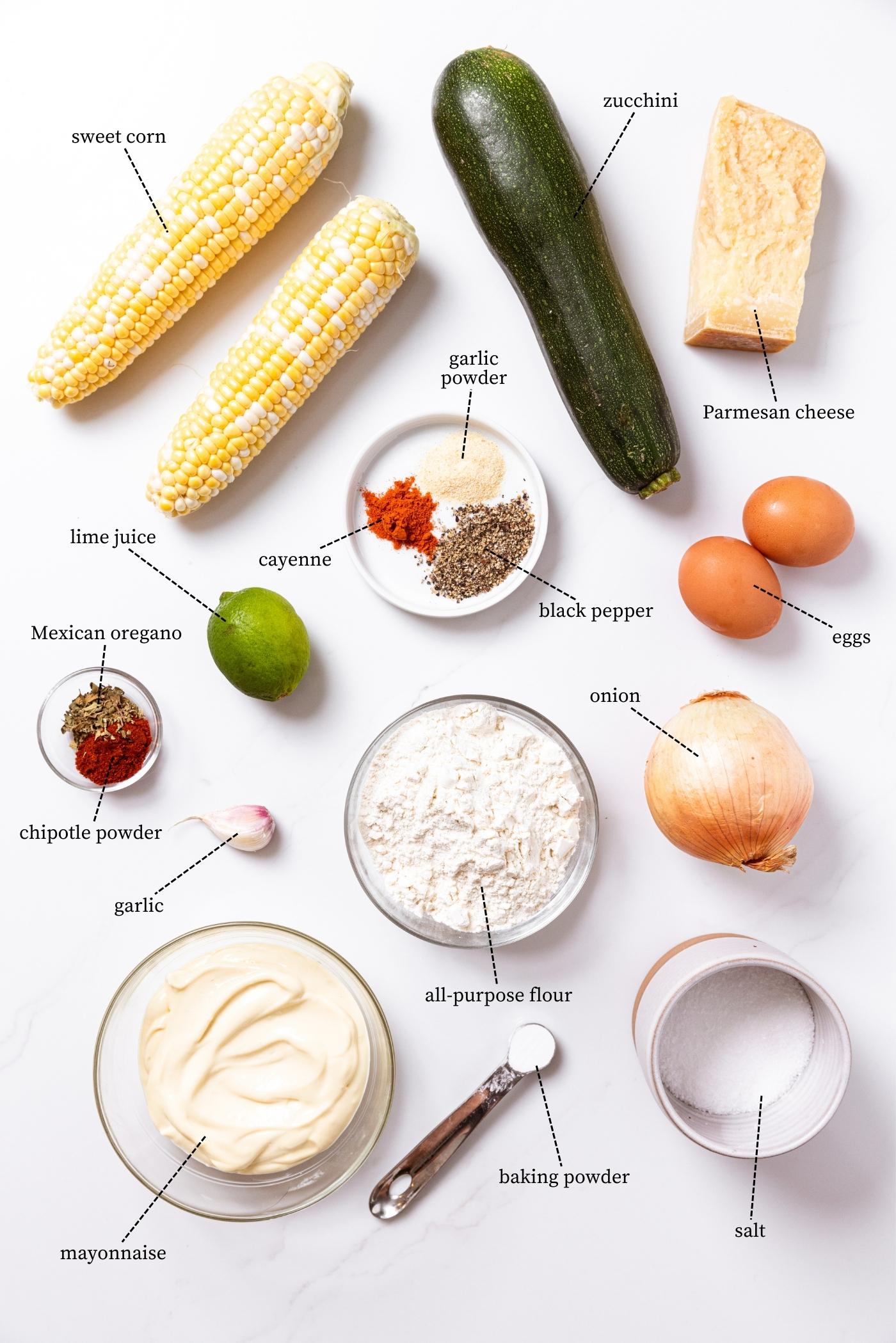 White countertop filled with ingredients needed to make zucchini and corn fritters including corn, zucchini, Parmesan cheese, spices, eggs, and more.