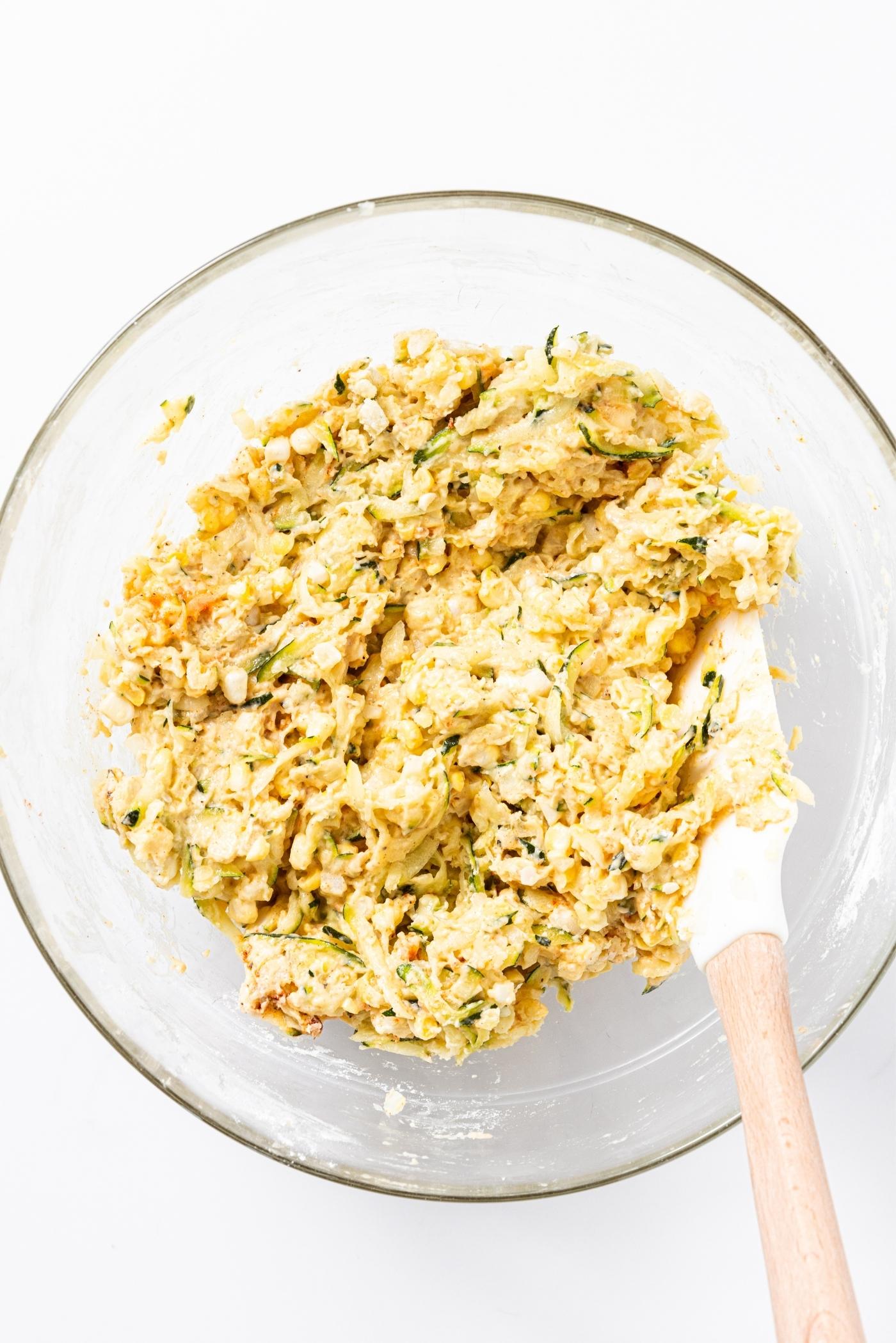 Glass bowl on white countertop with mixture of fritter batter with whisk resting on side of bowl.
