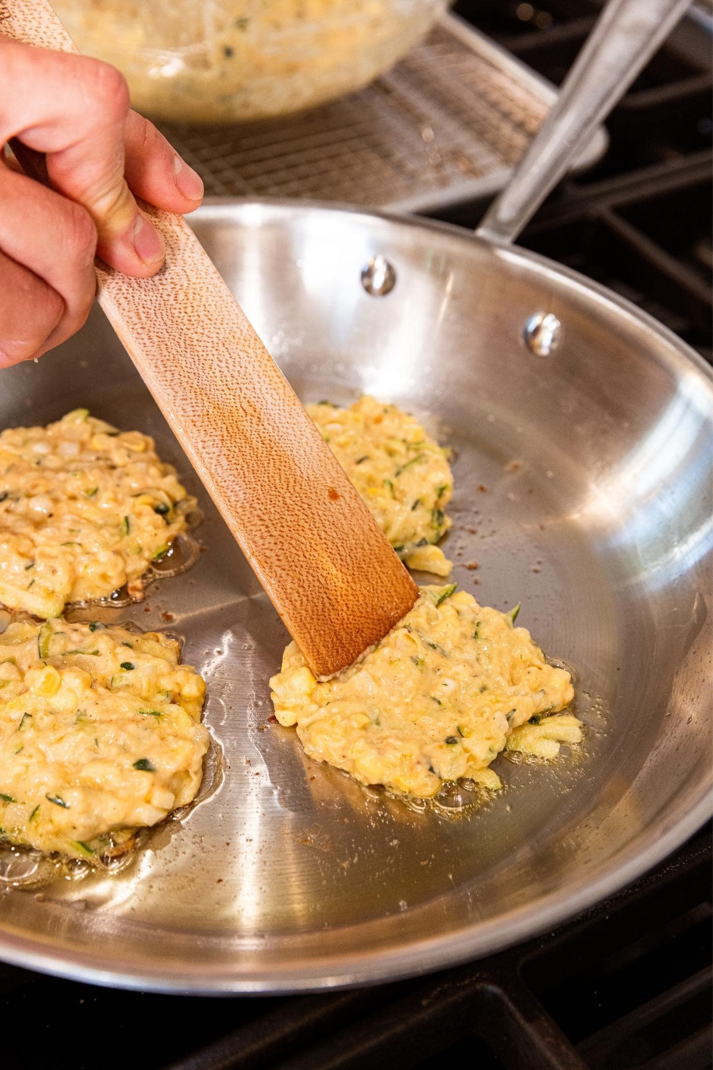 Hand pressing out fritter batter with wooden spatula on metal skillet on stove.