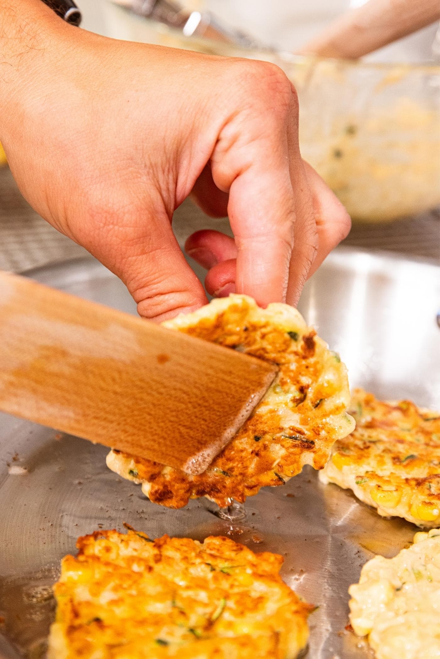 Hand using wooden spatula to flip corn fritter in metal skillet on stove.