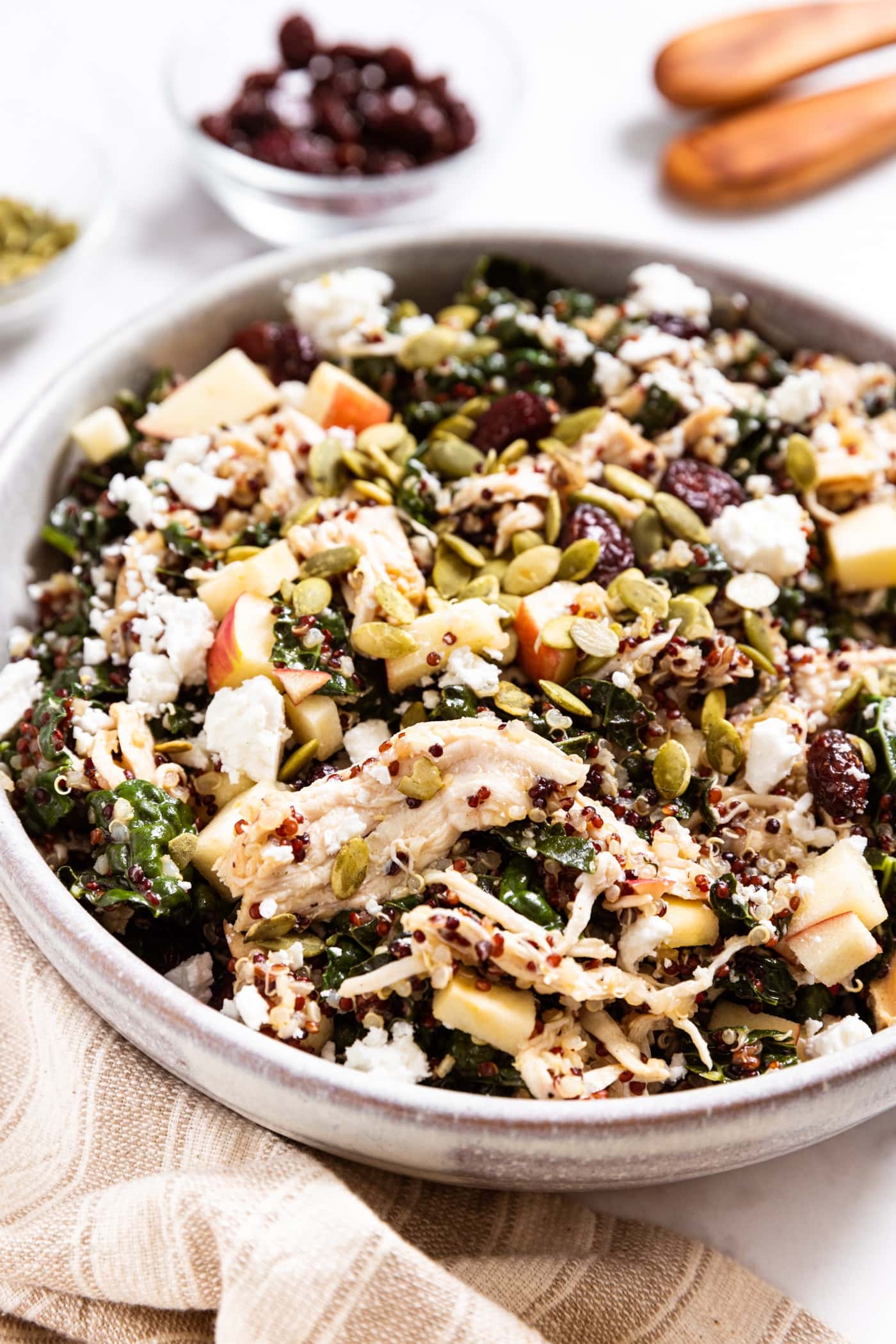 Stoneware bowl sitting on marble surface filled with quinoa kale salad topped with shredded chicken, pieces of apple, cranberries, pumpkin seeds, and feta cheese.