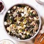 Large stoneware bowl filled with apple cider kale salad topped with pumpkin seeds, feta cheese, and cranberries all on white surface.