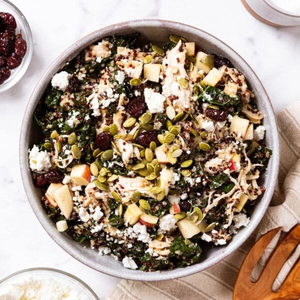 Large stoneware bowl filled with apple cider kale salad topped with pumpkin seeds, feta cheese, and cranberries all on white surface.