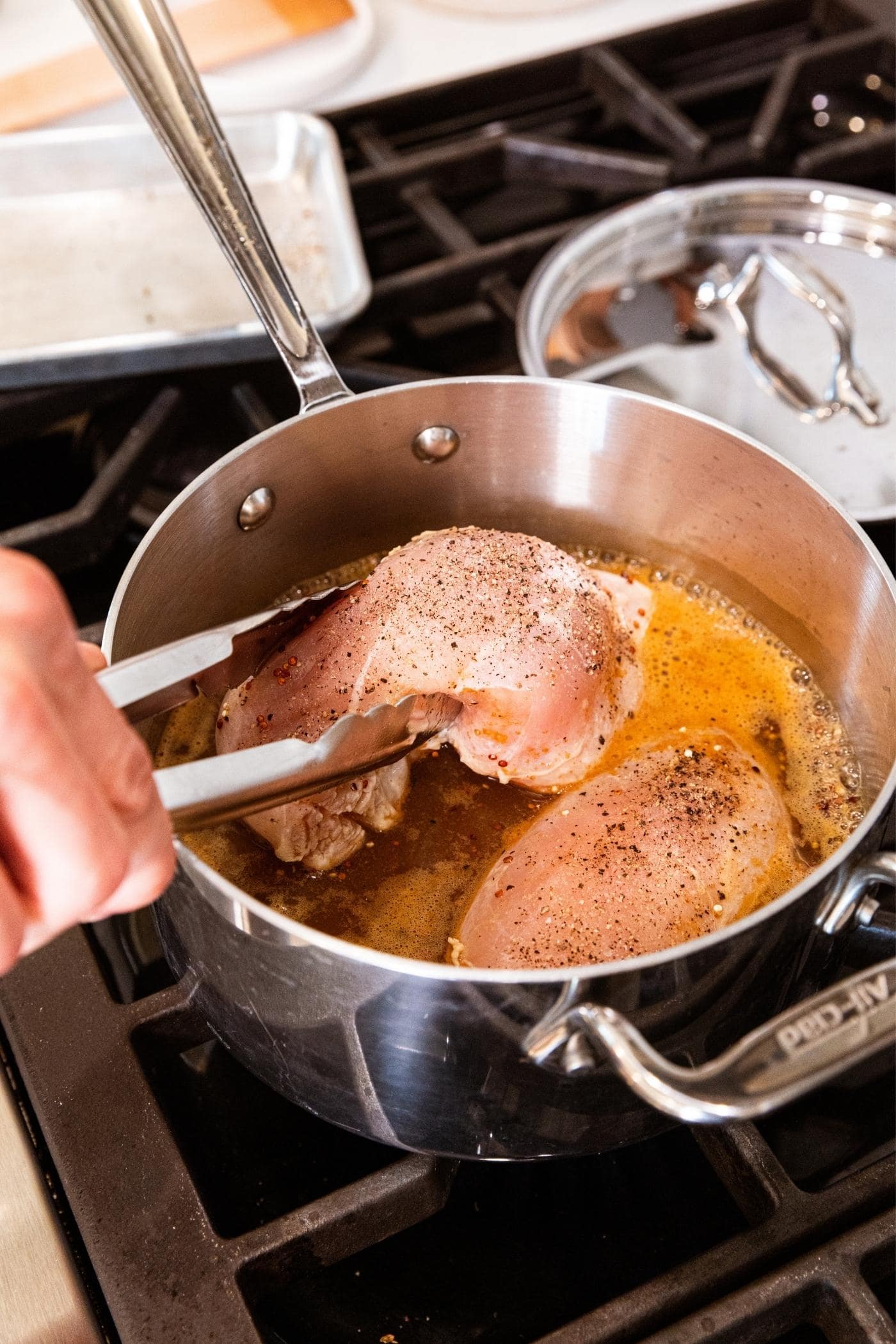 Hand putting raw chicken breasts into simmering apple cider liquid in kettle on stovetop.