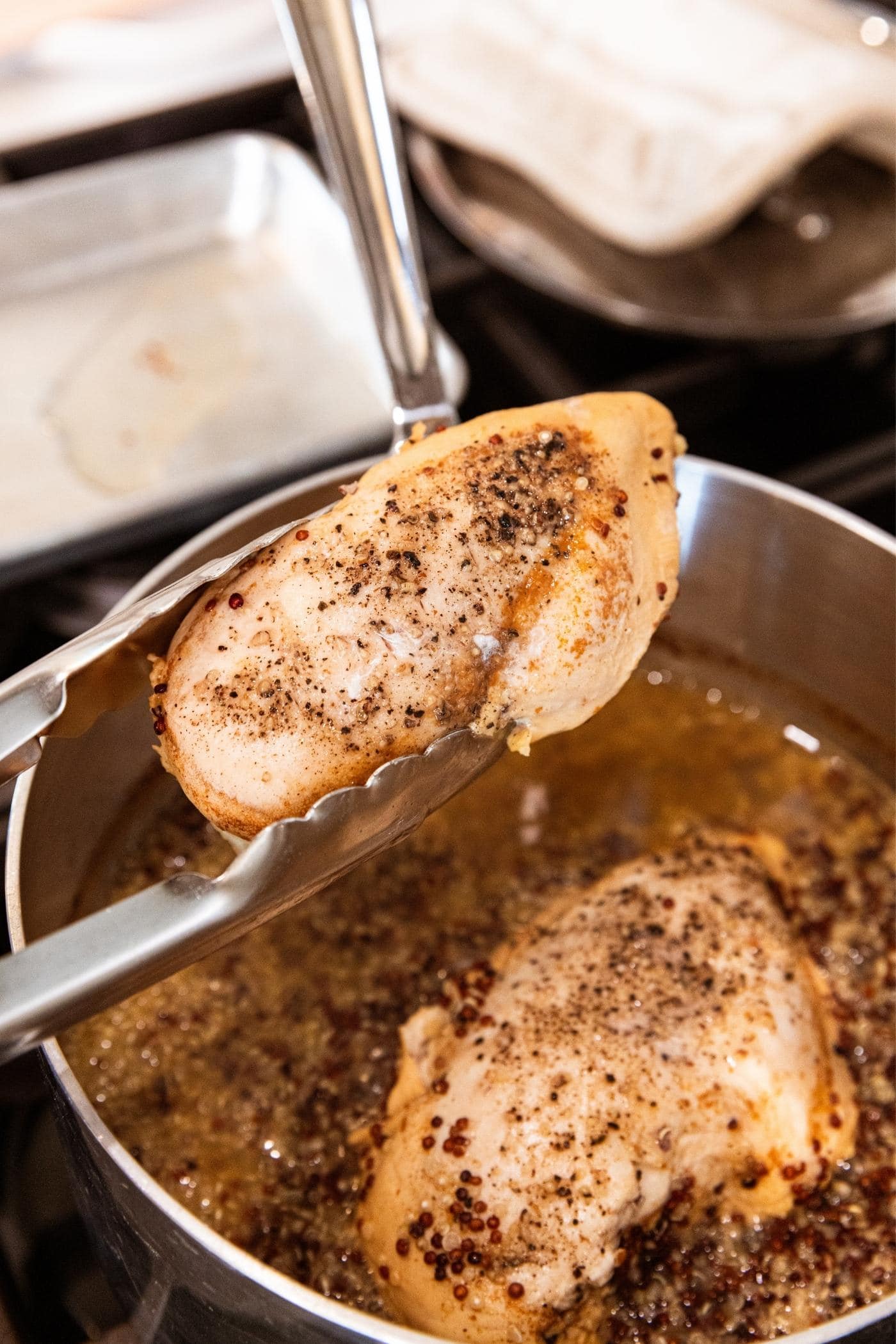 Hand using tongs to pull cooked chicken breast out of apple cider and quinoa liquid from kettle on stovetop.