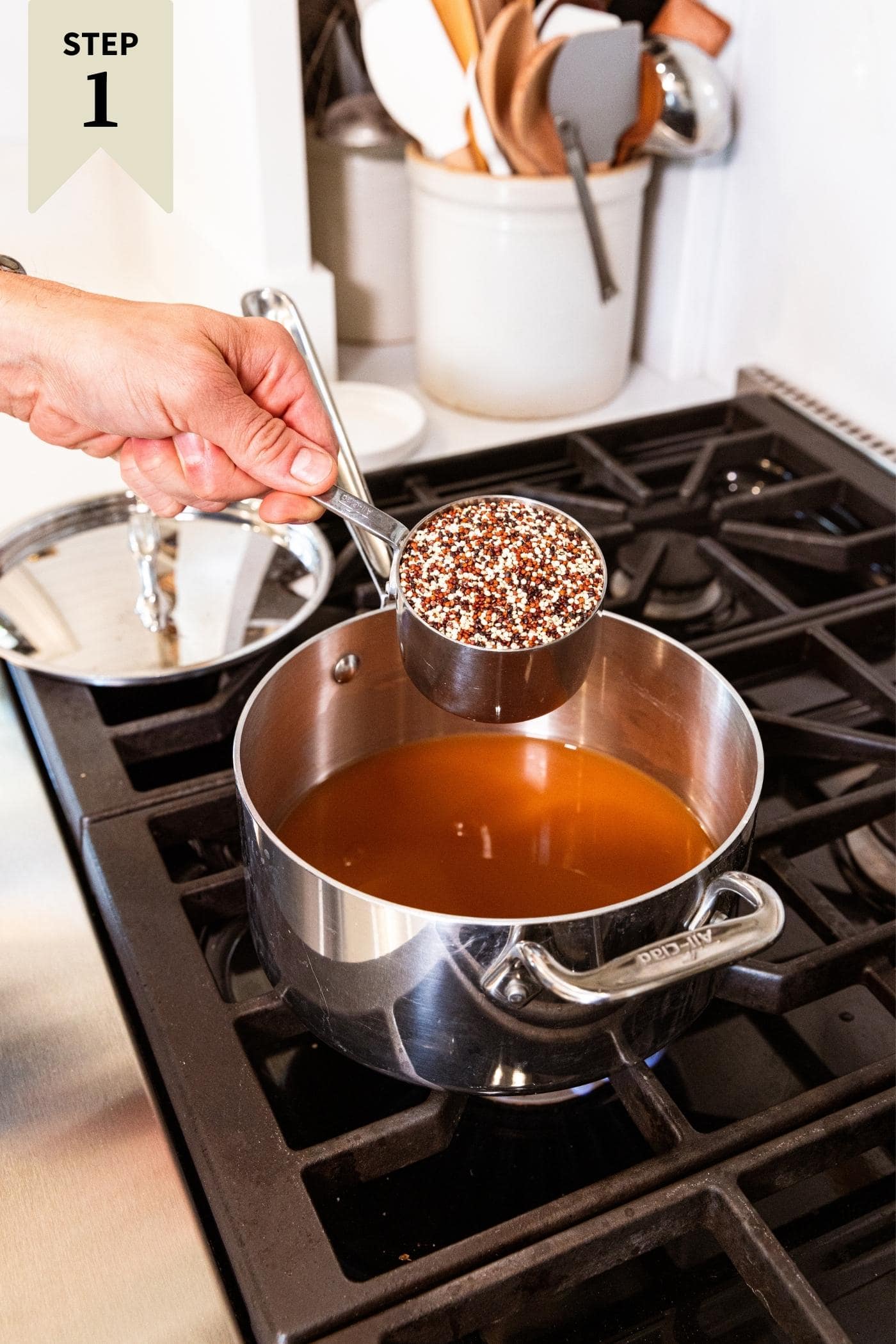 Hand pouring measuring cup filled with multicolored quinoa into kettle on stove filled with apple cider.