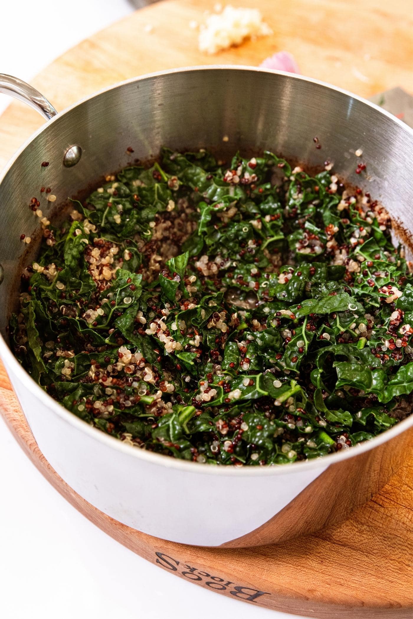 Metal kettle sitting on wood cutting board with the inside filled with cooked kale and dark green quinoa.