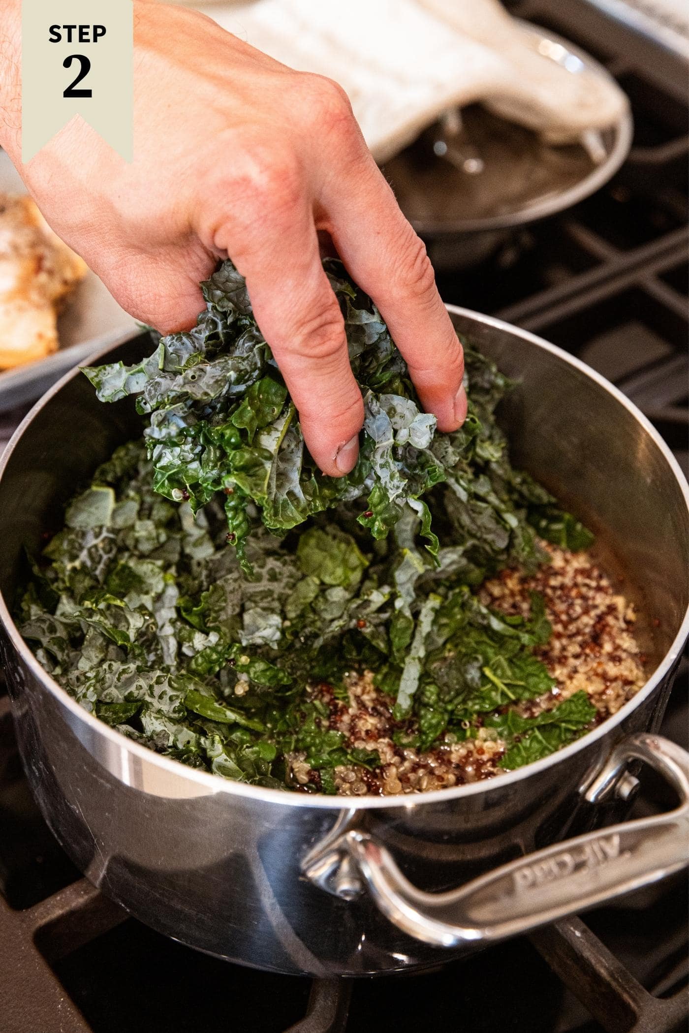 Hand placing chopped pieces of kale into metal pot on stovetop filled with cooked quinoa.