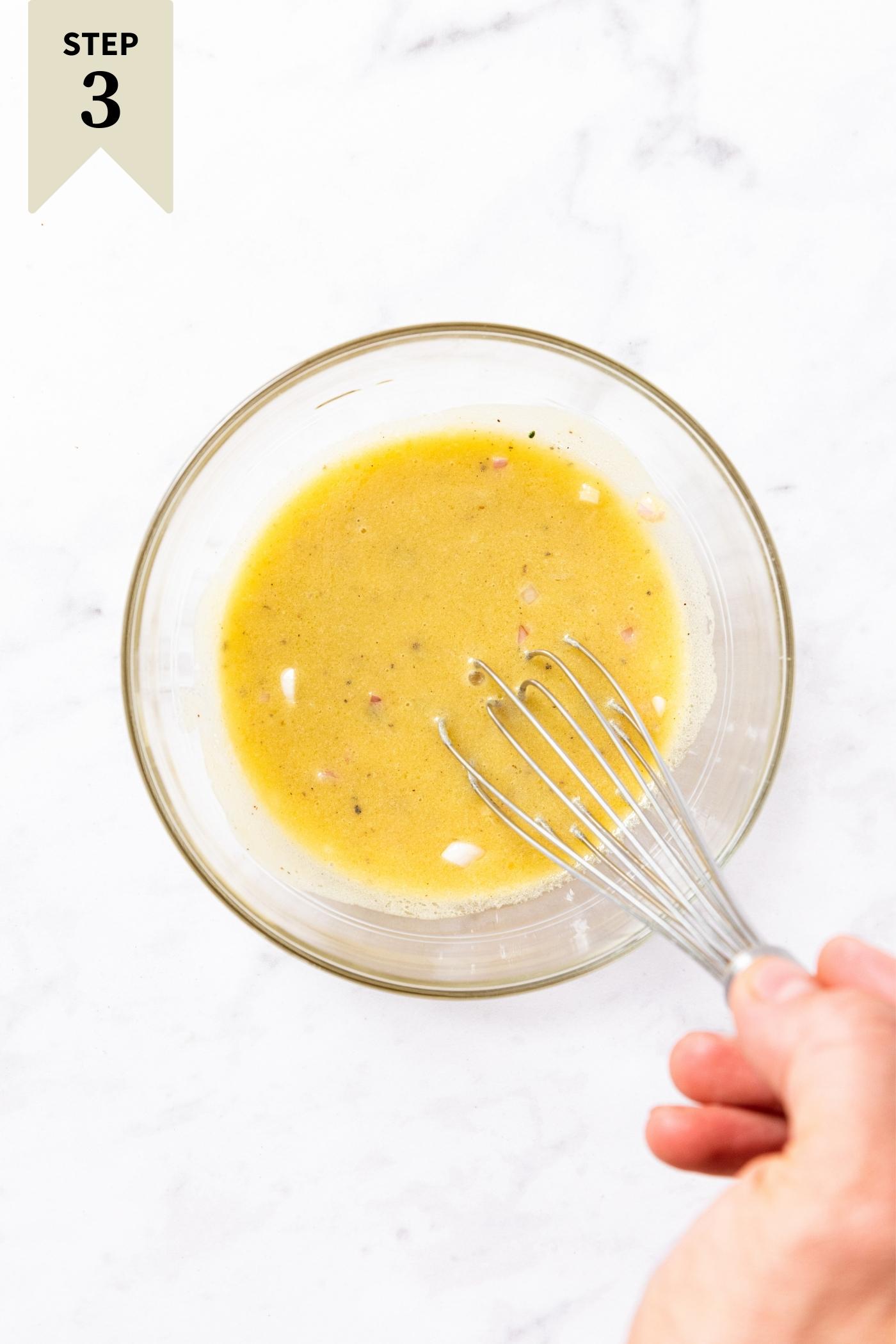 Glass bowl sitting on marble surface with hand using whisk to stir together the yellow contents.