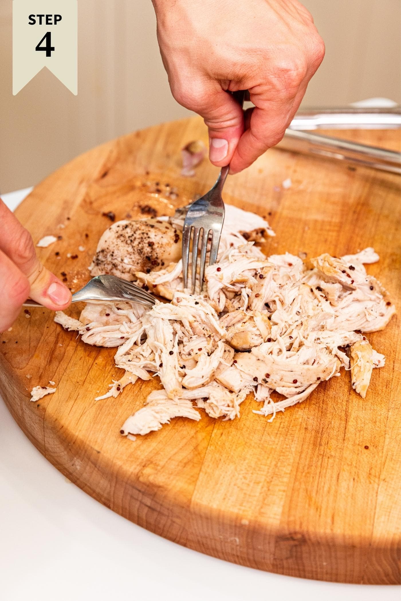 Two hands using two forks pulling apart pieces of poached chicken on wood cutting board.