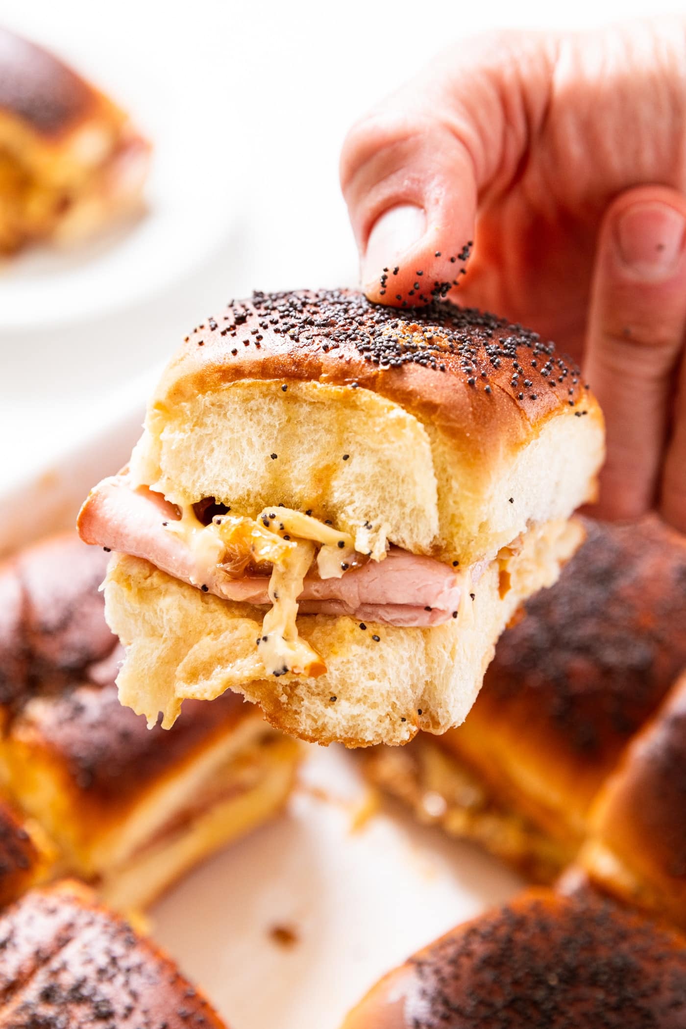 Hand holding a ham and cheese slider above the rest of the sandwiches in a white baking dish below.