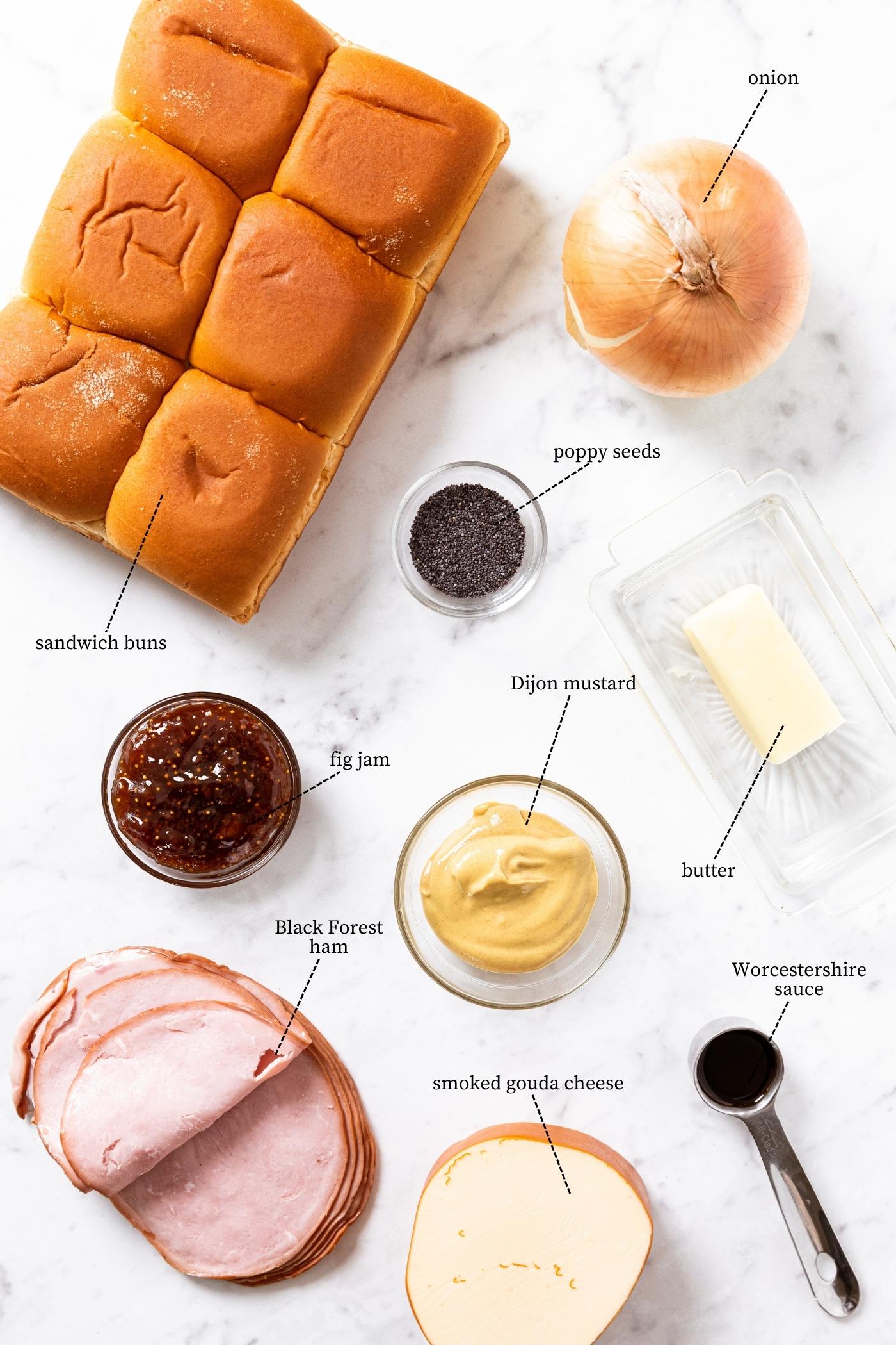 Marble countertop with all the ingredients needed to make slider sandwiches including small sandwich buns, ham, mustard, gouda cheese, fig jam, and more.