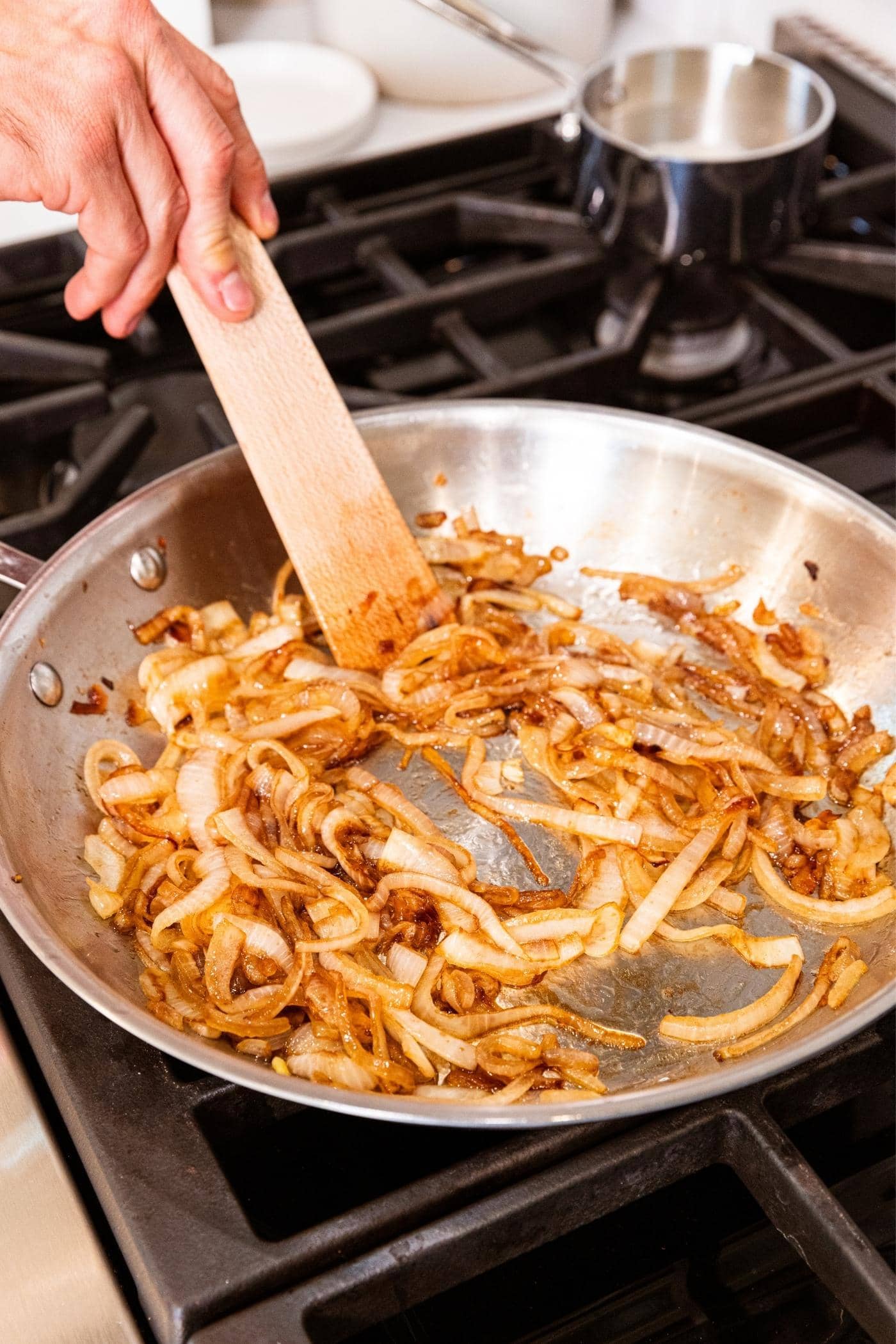 Hand pushing around brown caramelized onions in metal skillet with wooden spatula.