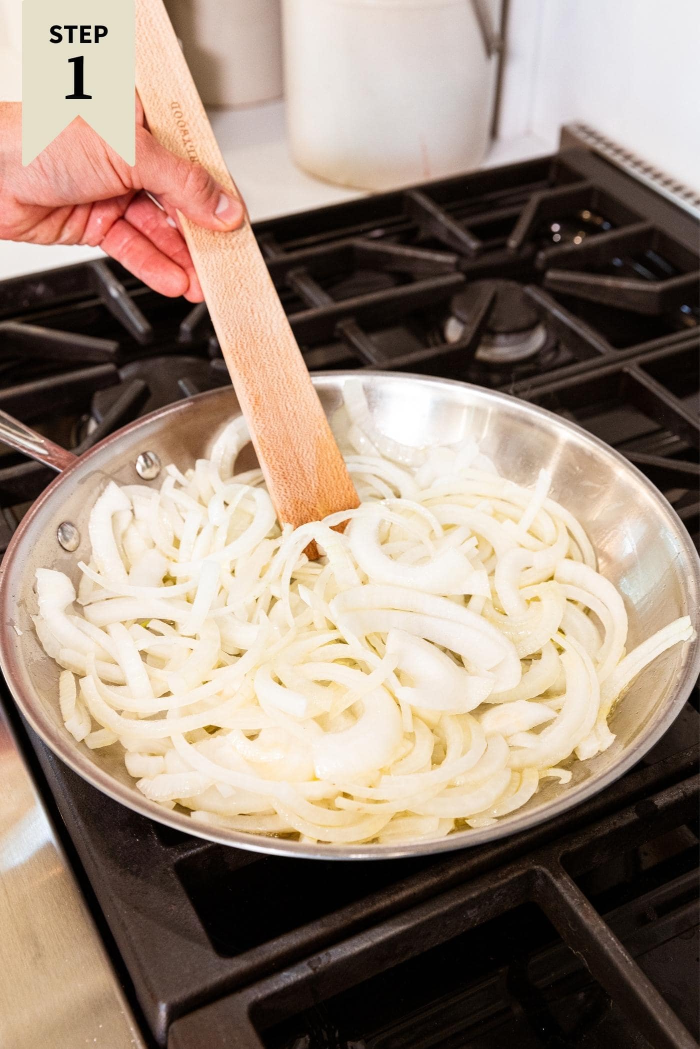 Hand holding wooden spatula pushing around raw onions in metal skillet on stovetop before they caramelized.