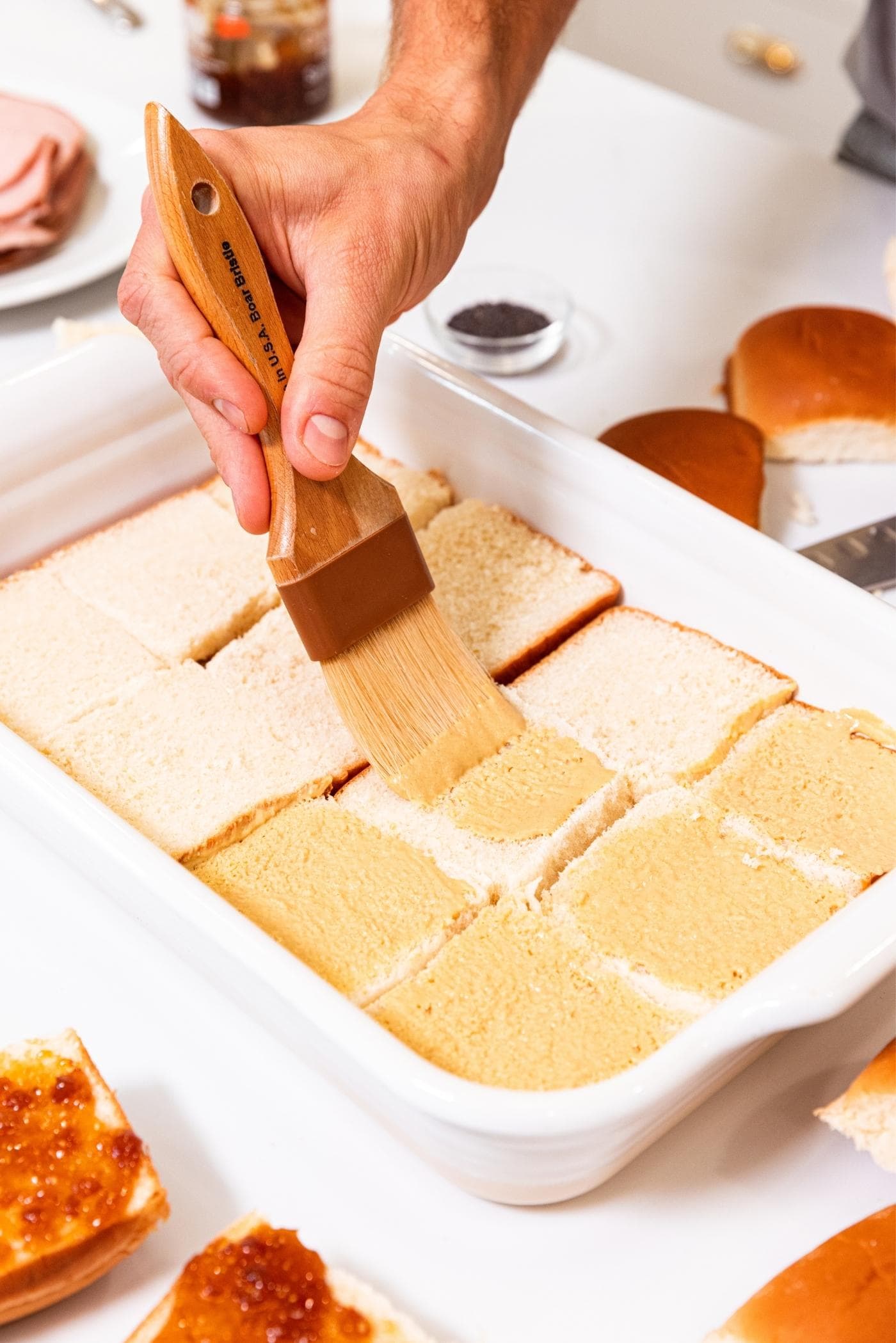 Hand using a brush to spread Dijon mustard onto bottom pieces of sandwich buns in white baking dish.
