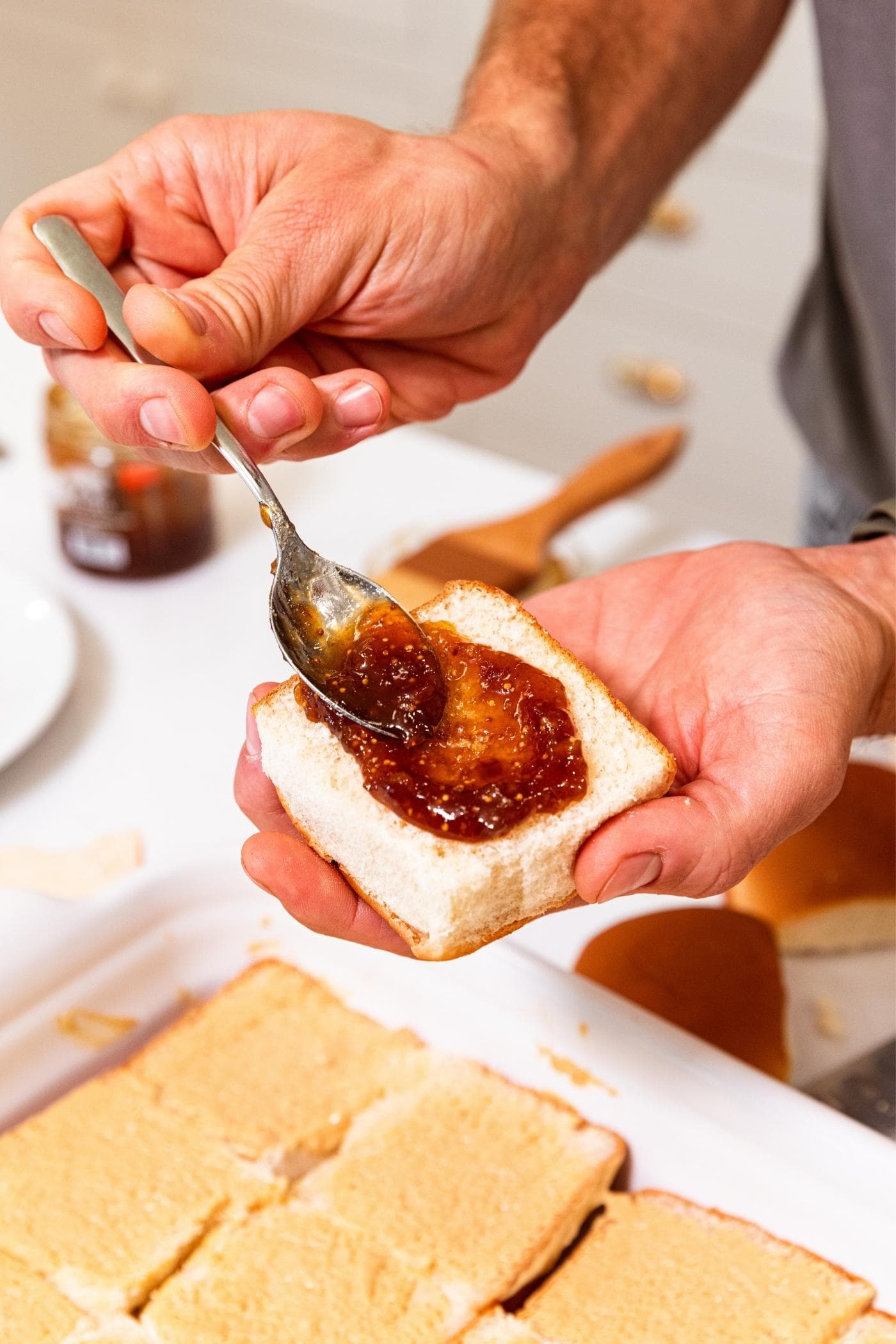 Hand holding the top of a sandwich bun spreading with dark purple colored fig jam with a spoon.