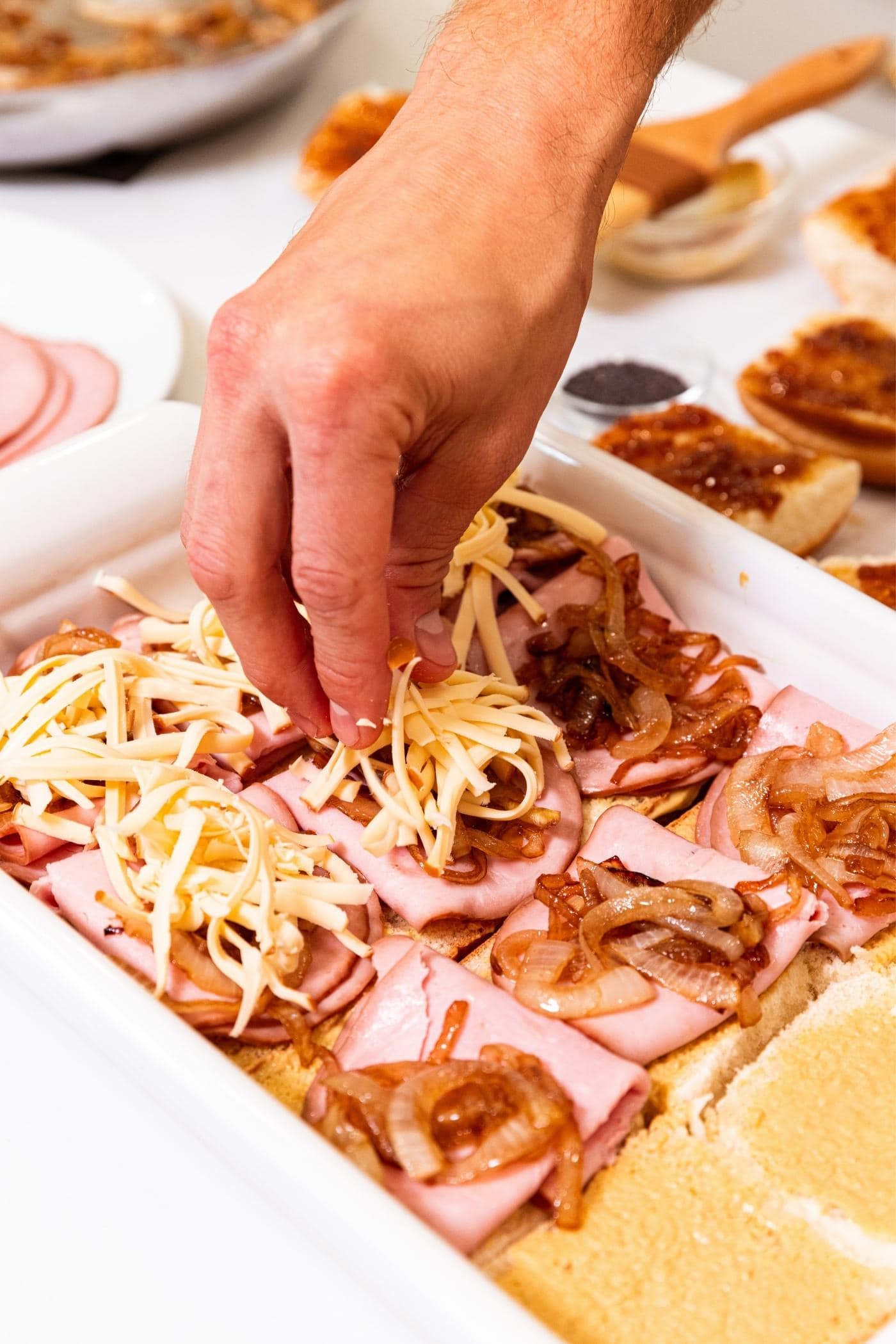 Hand dropping shredded cheese on top of sandwiches being assembled in a baking dish.