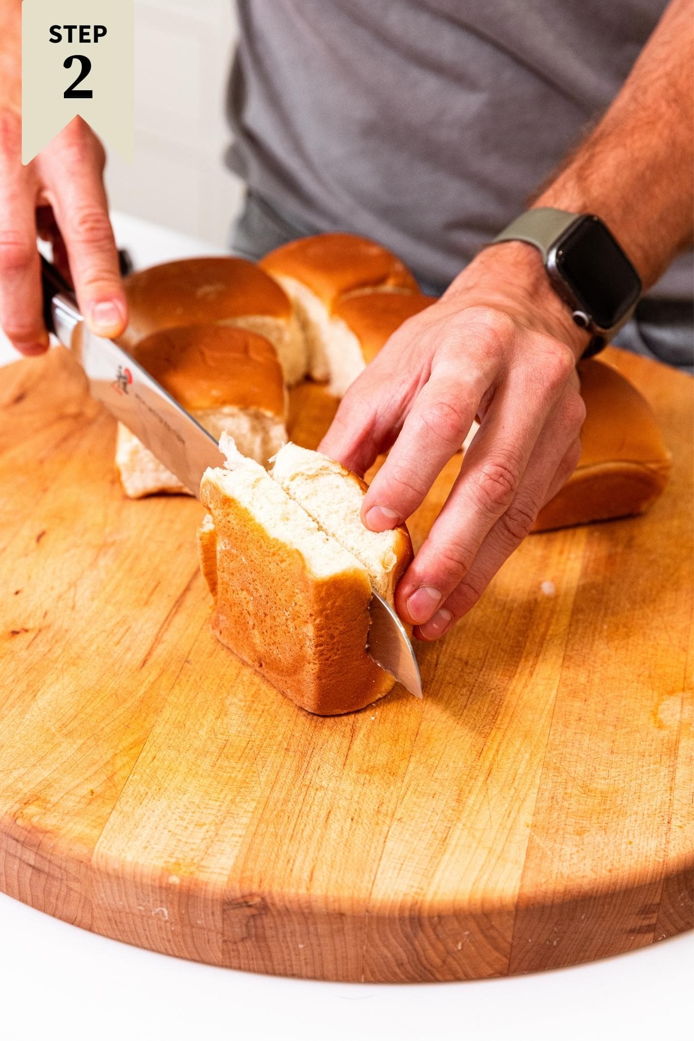 Hand using knife to slice a small sandwich bun in half on wood cutting board with more sandwich buns in half.