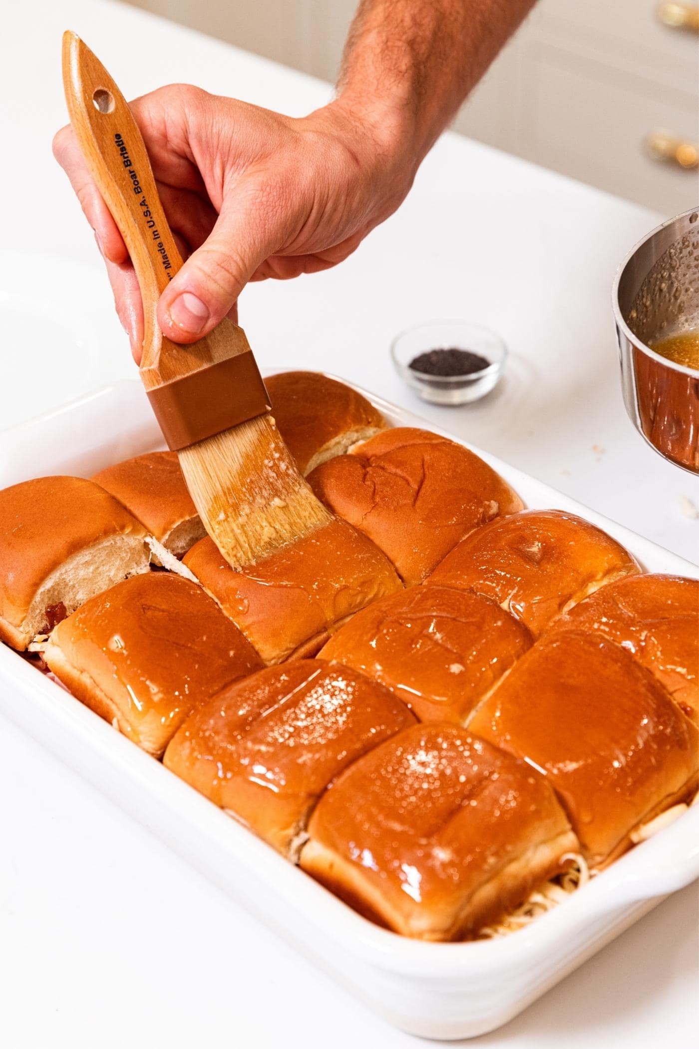 Hand brushing melted butter on top of sandwich buns sitting in white baking dish on white countertop.