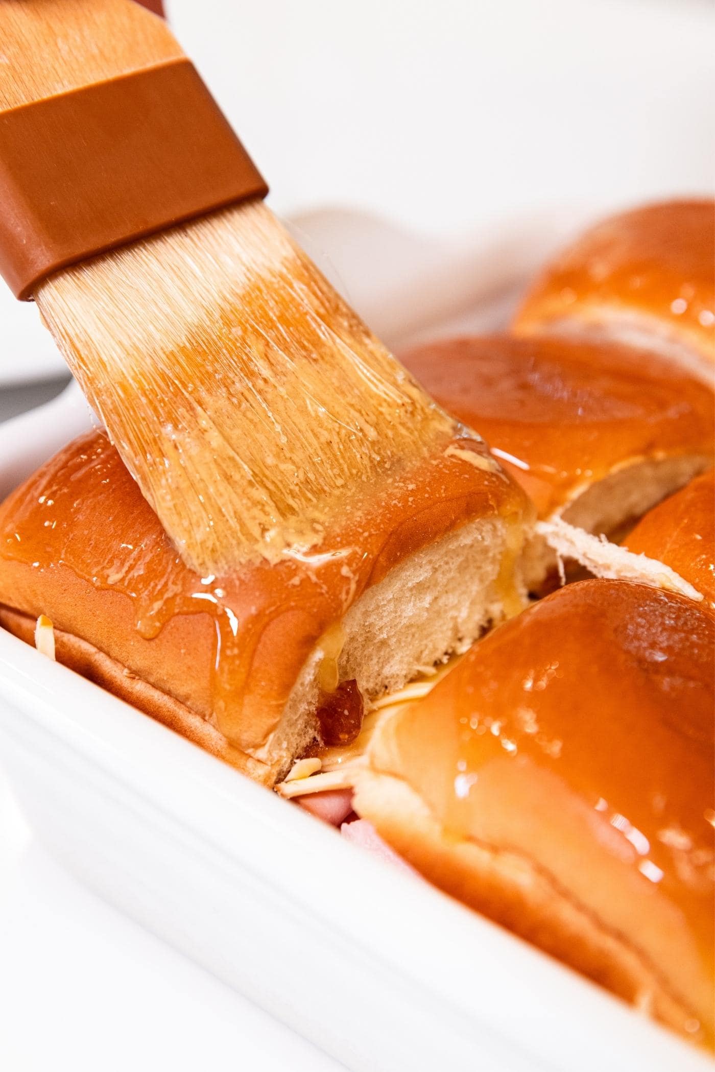 Brush spreading on melted butter on top of sandwich bun in white baking dish.