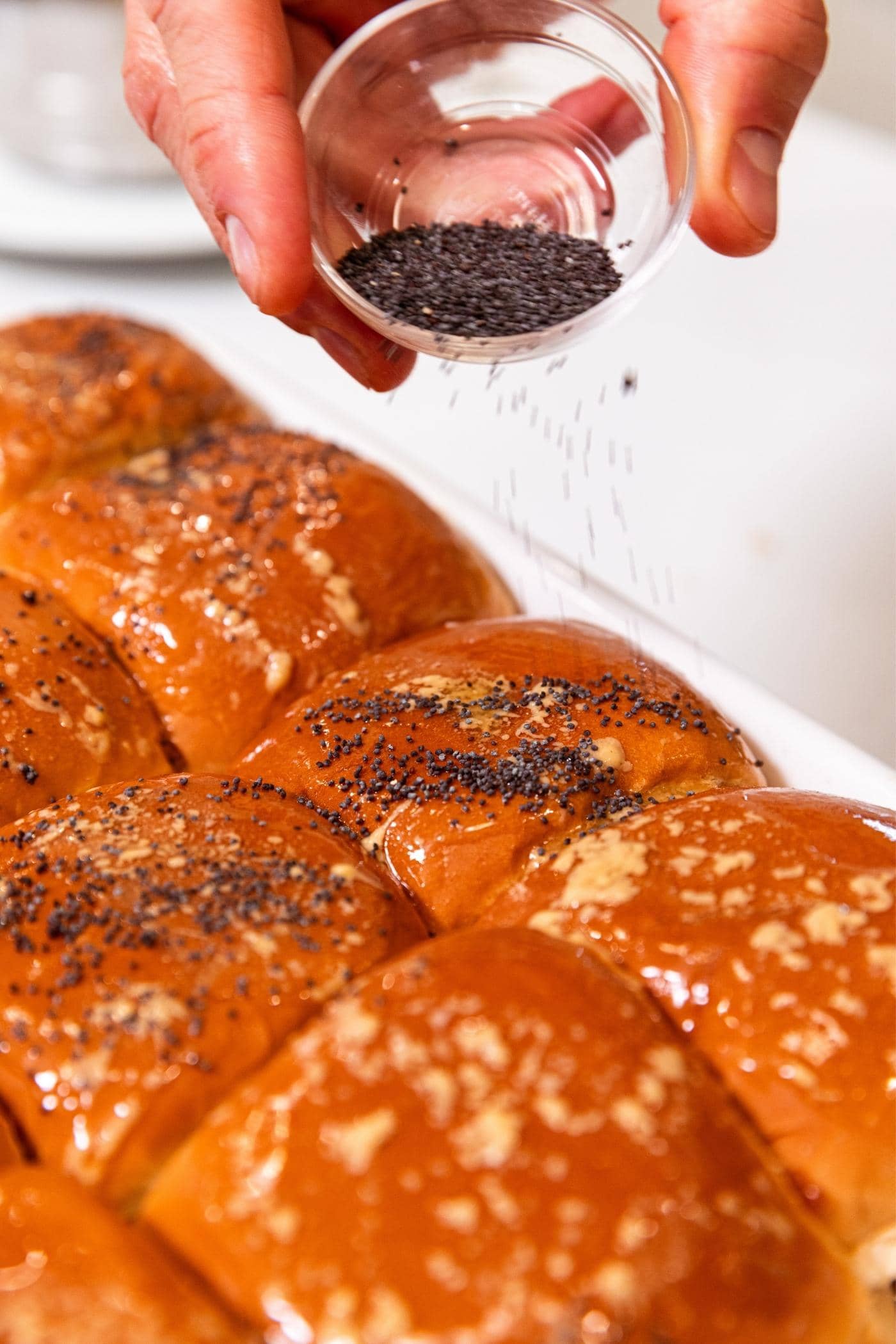 Hand holding small glass bowl filled with poppy seeds sprinkling them on top of sandwich bun brushed with melted butter in white baking dish.