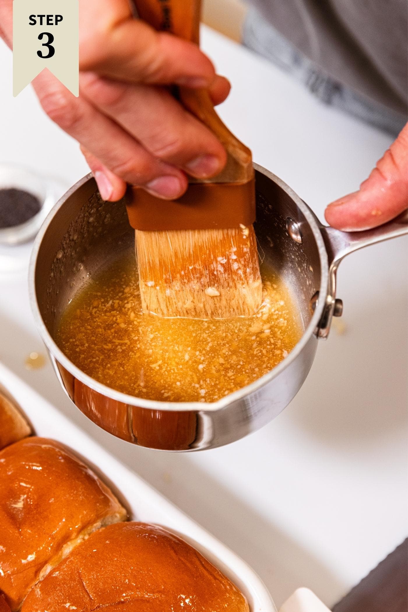 Hand holding small pot with melted butter inside and brush dipping inside before spreading on top of a bun.