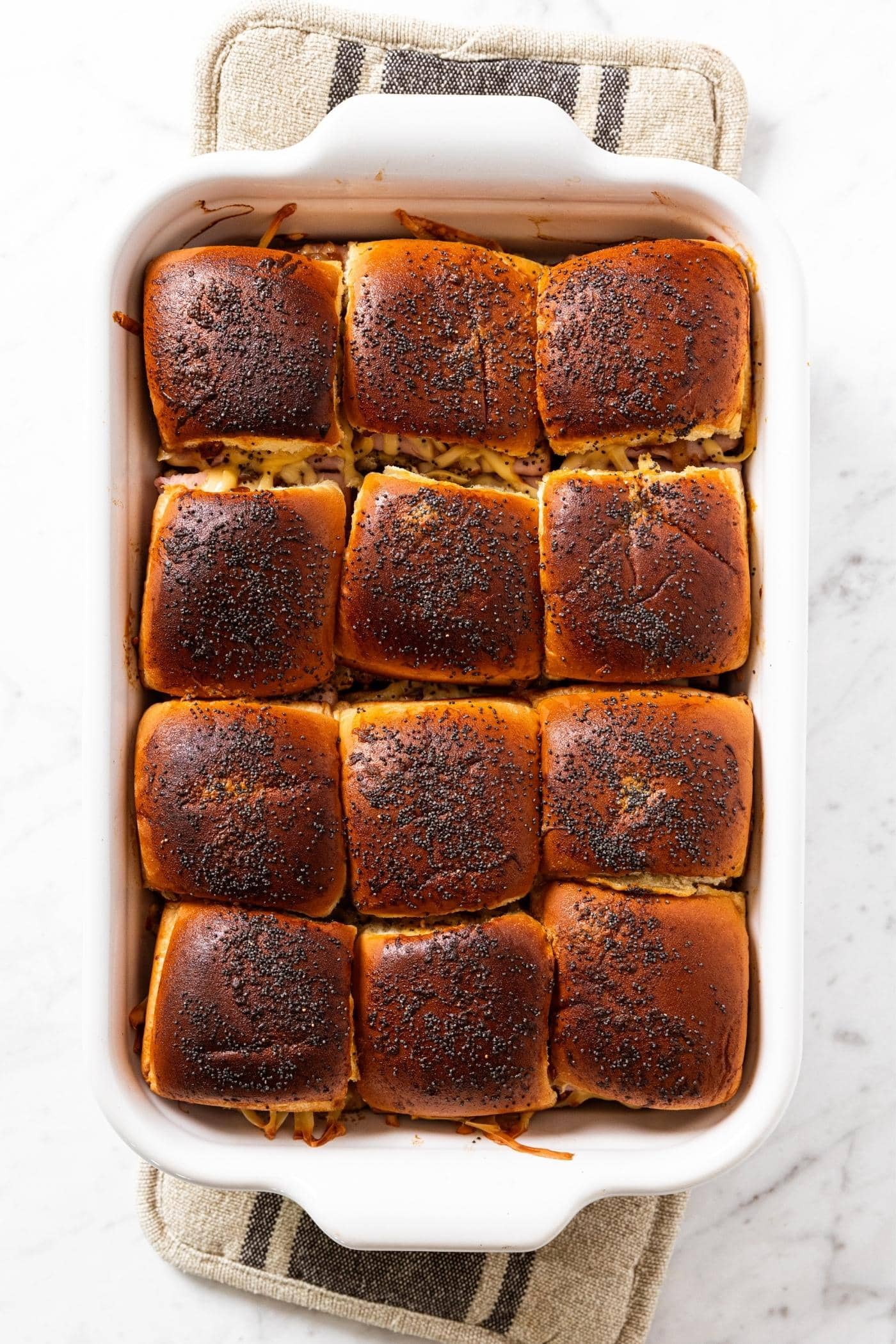 Baked sandwich buns in white baking dish sitting on marble countertop with hot pads underneath with tops of sandwich buns browned by the baking.