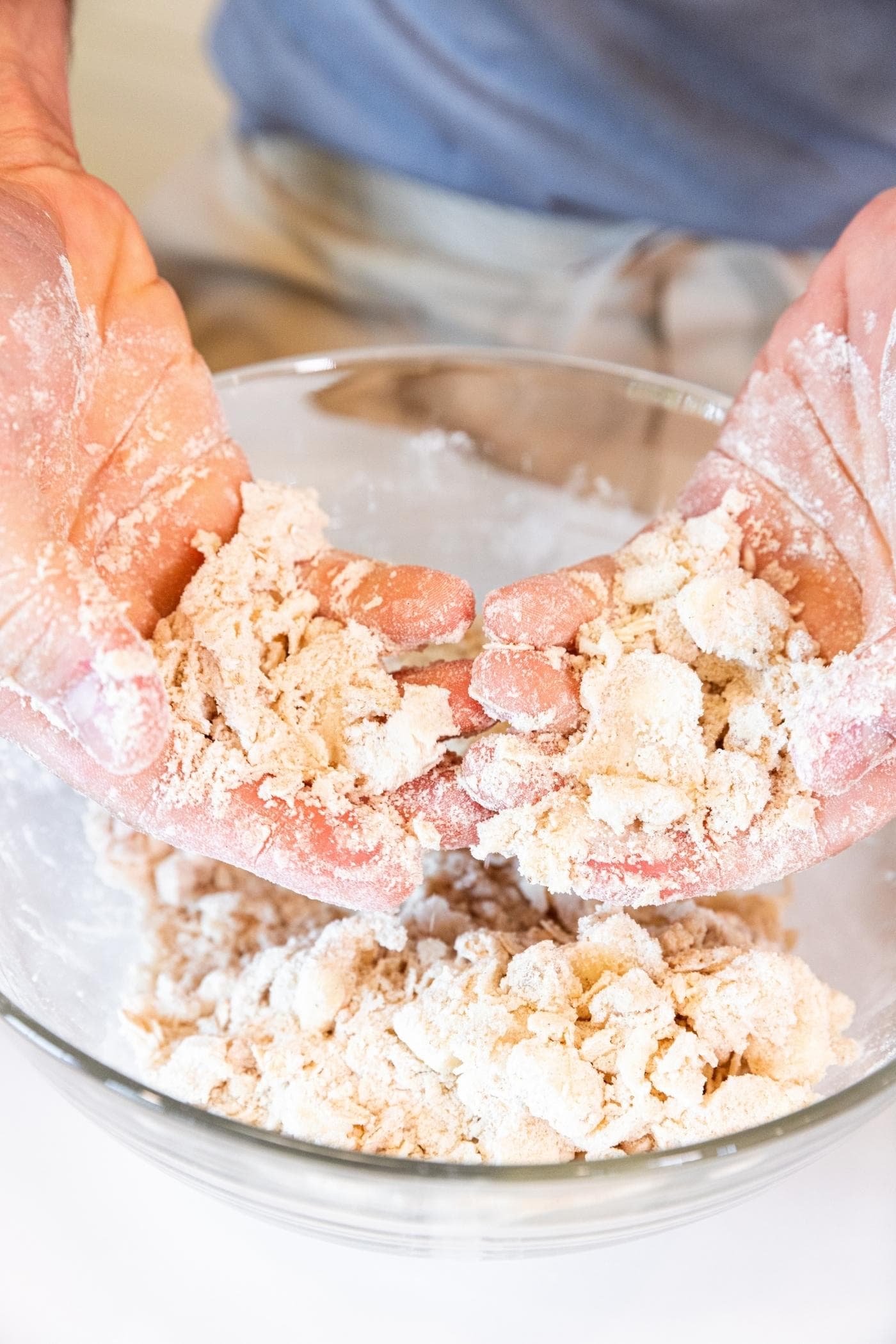 Two hand showing crushed up butter and flour forming a dough in a glass bowl on white surface.