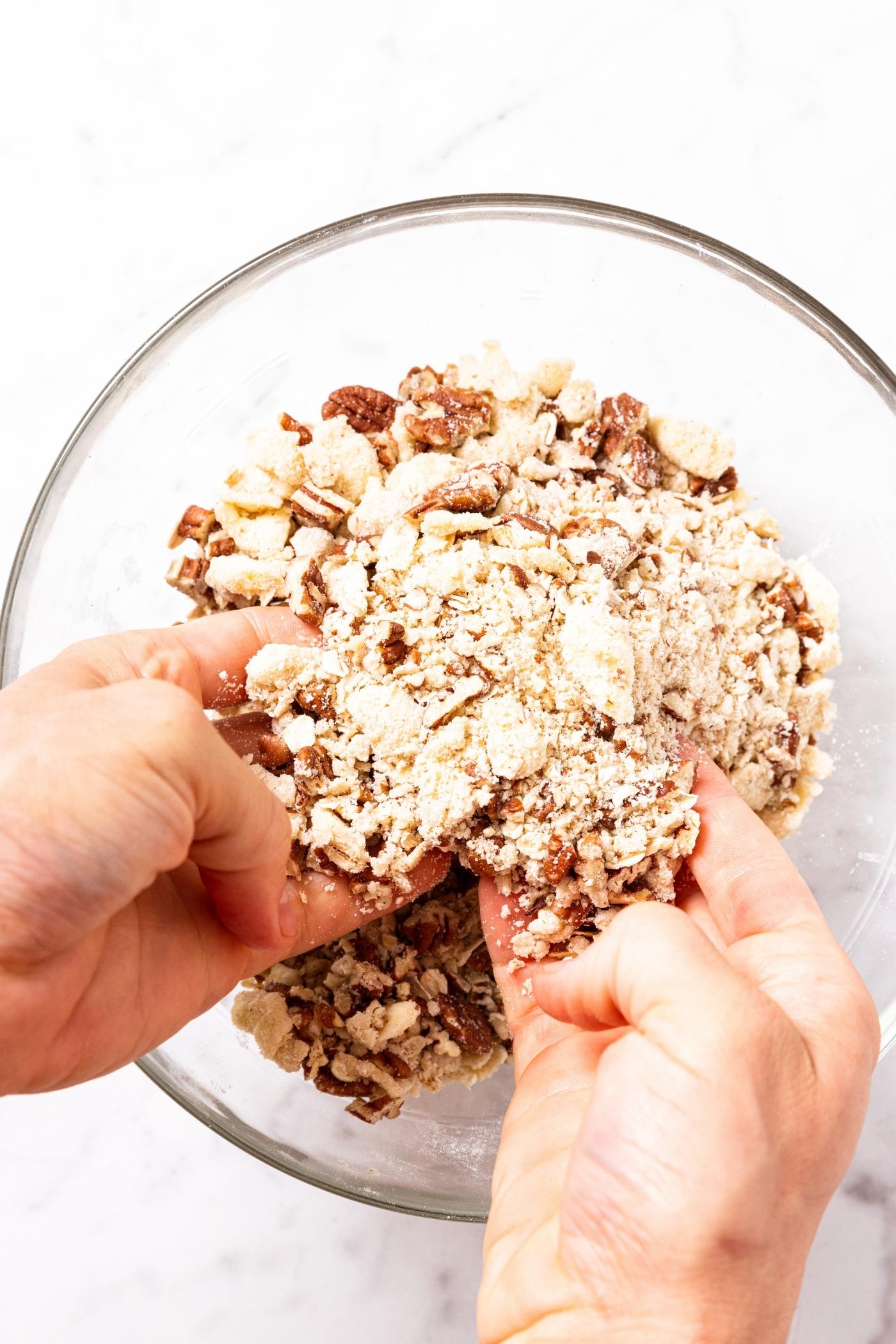 Two hands holding streusel topping filled with chopped pecans in glass bowl on marble surface.