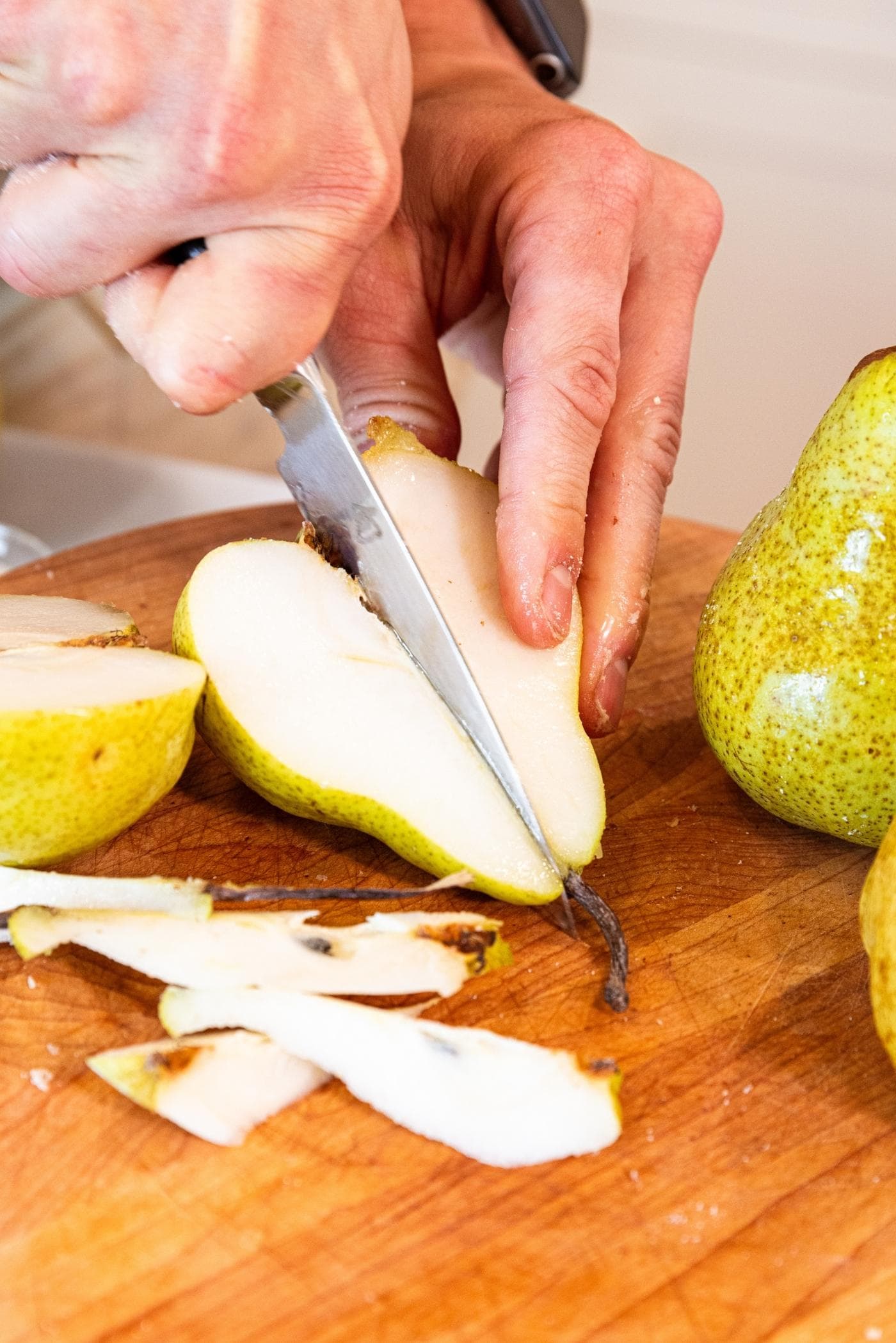 Hand slicing halved pear in half again on wood cutting board with more pears all around.