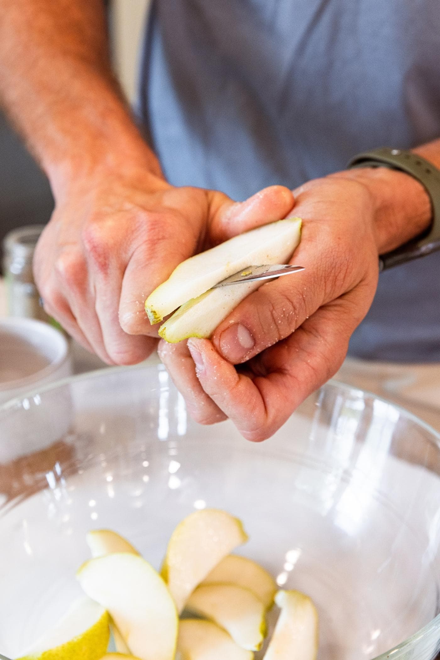 Two hands slicing pear with skin still on into a glass bowl with more pears slices already inside.