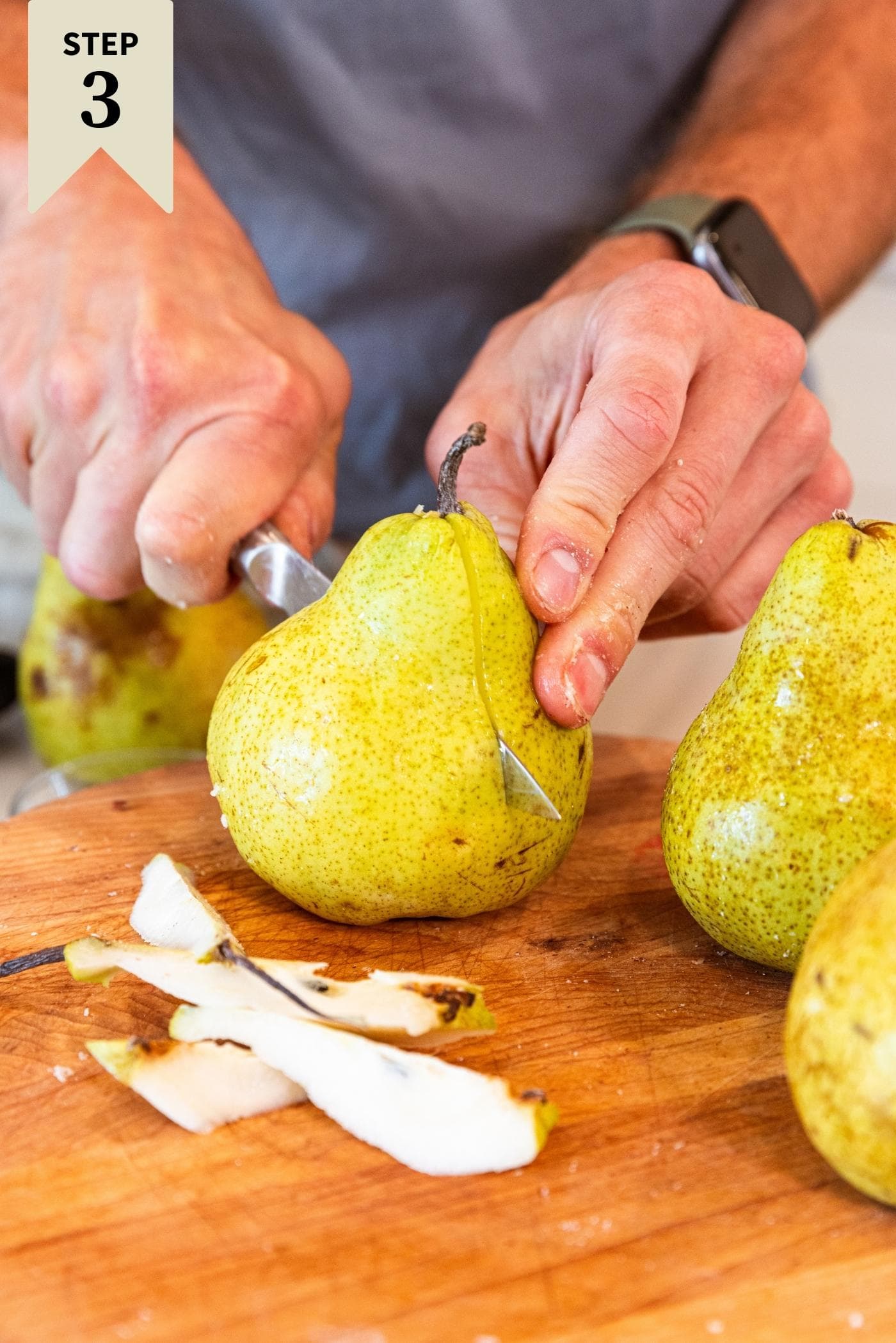 Hand slicing green colored pear in half on wood cutting board with more pears around.