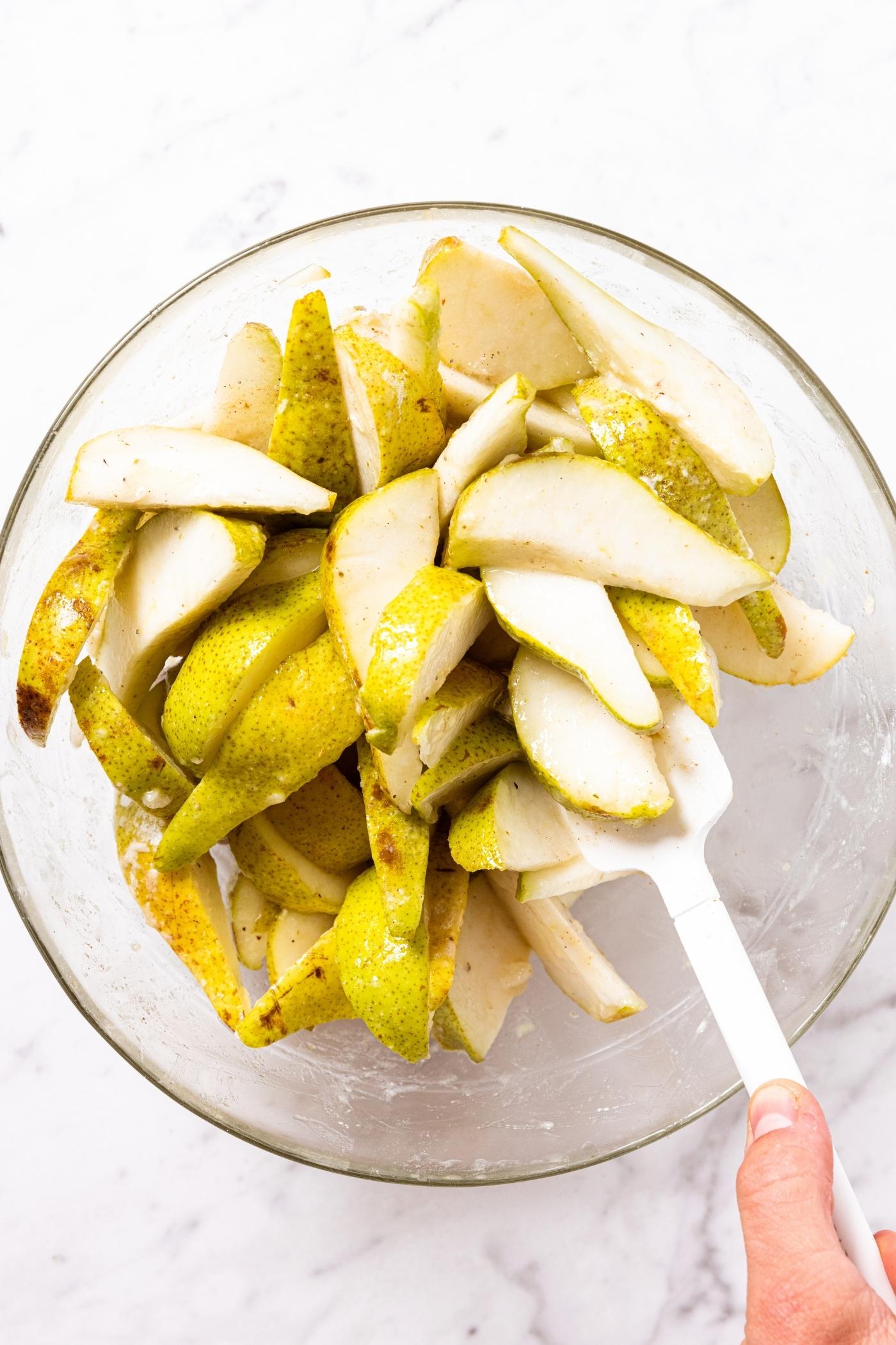 Glass bowl filled with slices of pears with white spatula stirring together all pears with spices sprinkled on top.