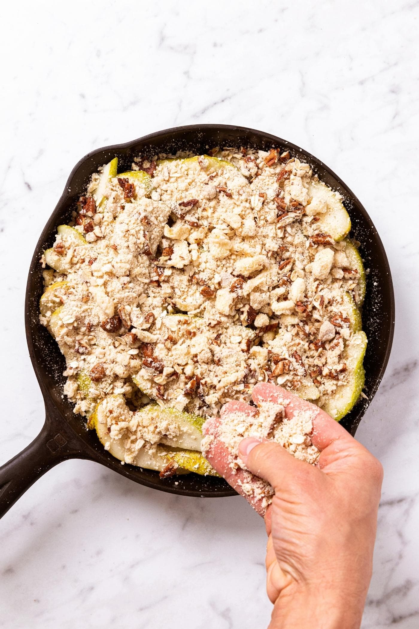 Hand sprinkling streusel topping all over sliced pears in black skillet on marble surface before baking.