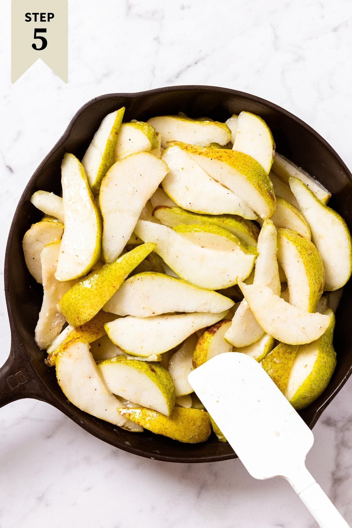 White spatula pushing around pears in black skillet on marble surface.