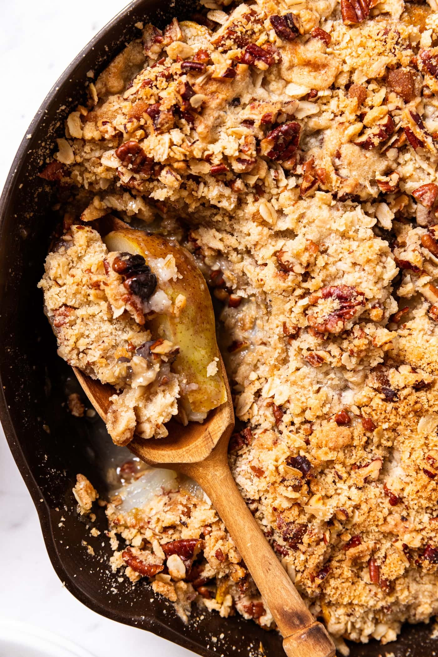 Black cast iron skillet filled with pear crisp with wooden spoon resting in pan filled with serving of crisp all on marble surface.