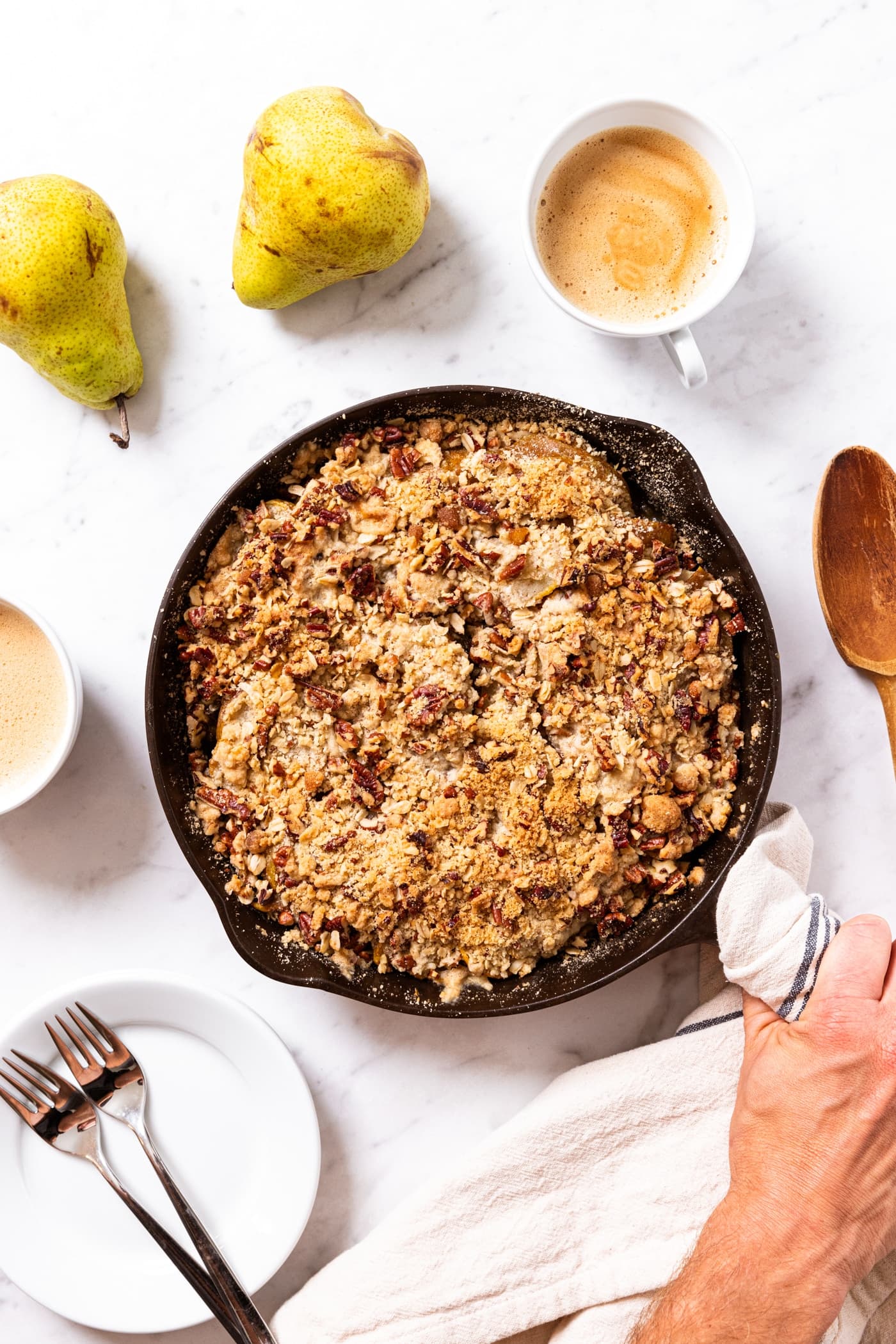 Hand holding black skillet filled with pear crisp with towel around hot handle with pears and cups of coffee also sitting on the marble surface.