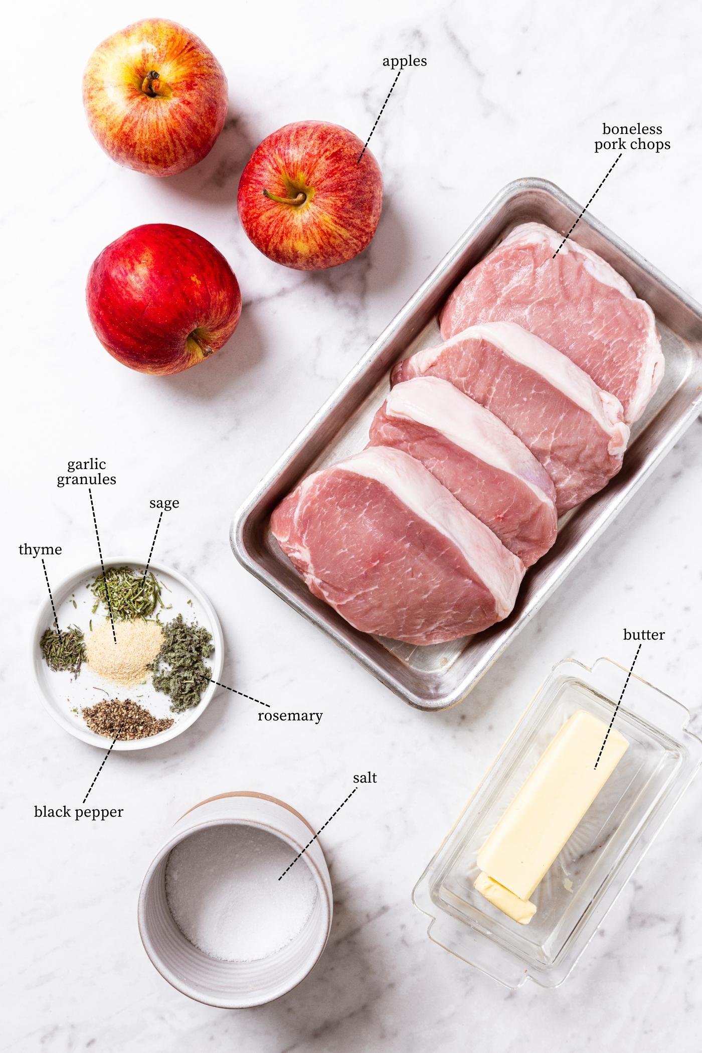Marble surface filled with all of the ingredients needed to make pork chops with quick applesauce including raw pork chops, salt, butter, apples, and spices.