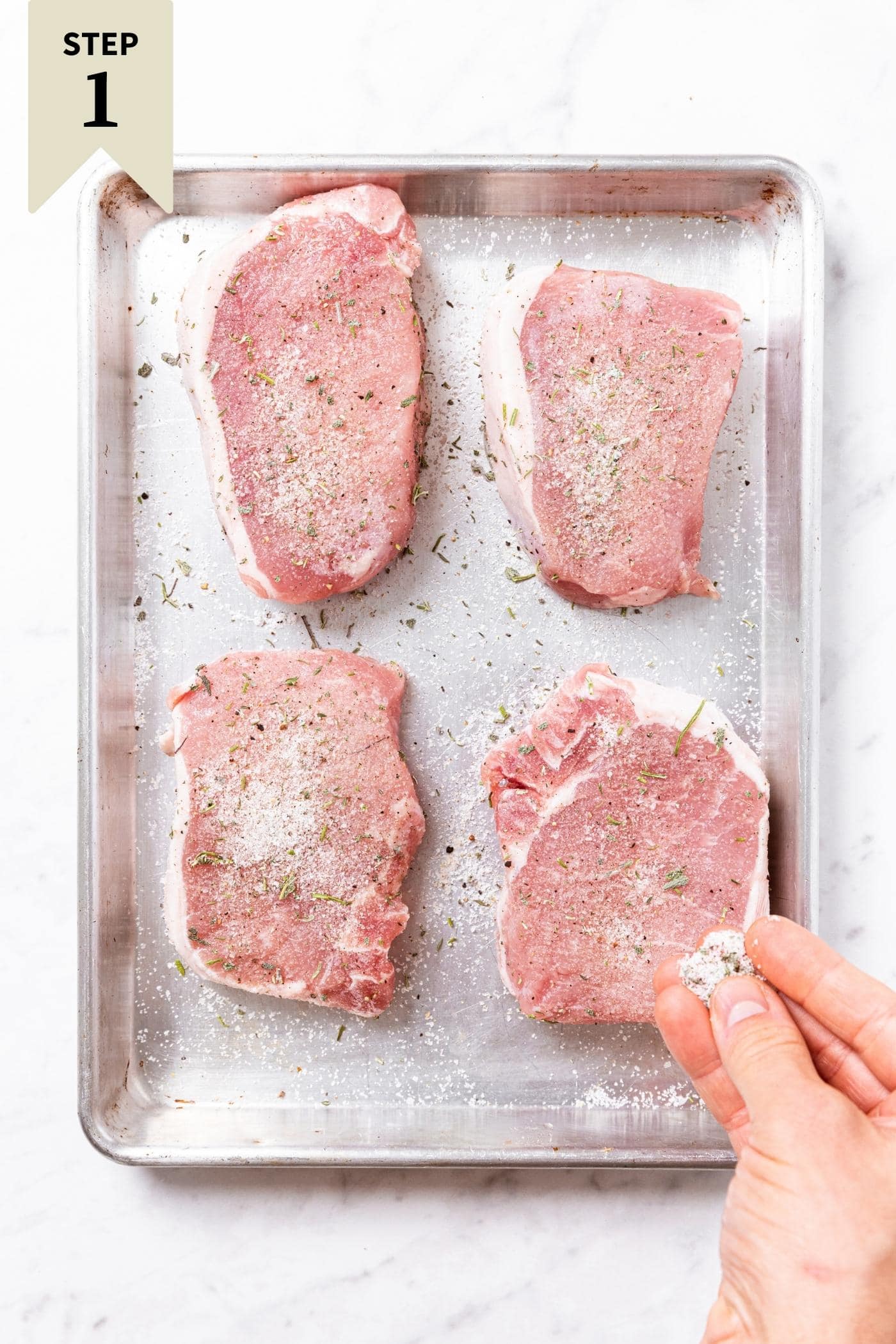 Hand sprinkling dry brine salt mixture on top of raw pork chops sitting on metal pan on marble surface.