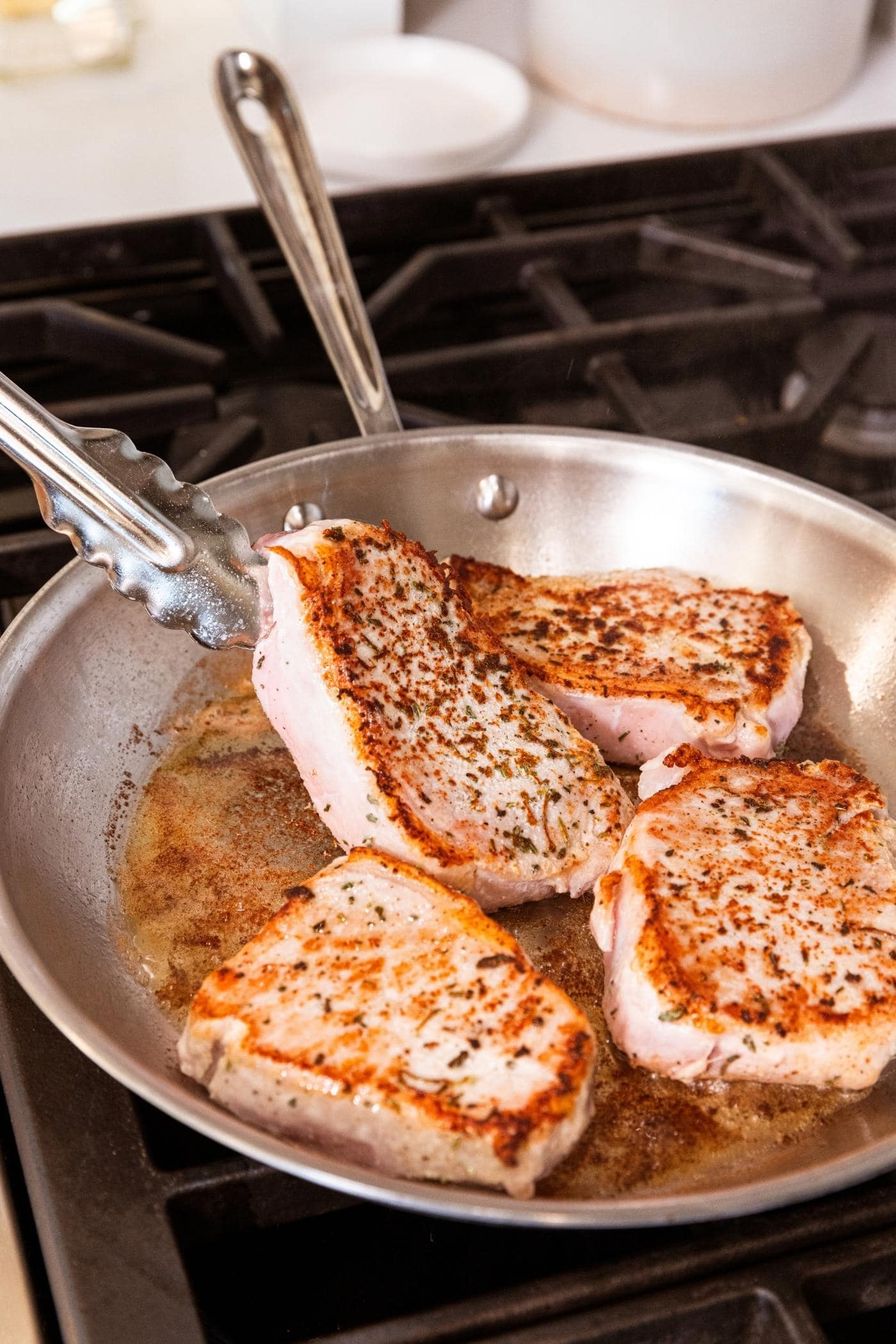 Tongs turning browned pork chop in metal skillet on stovetop with pan juices collected in pan.